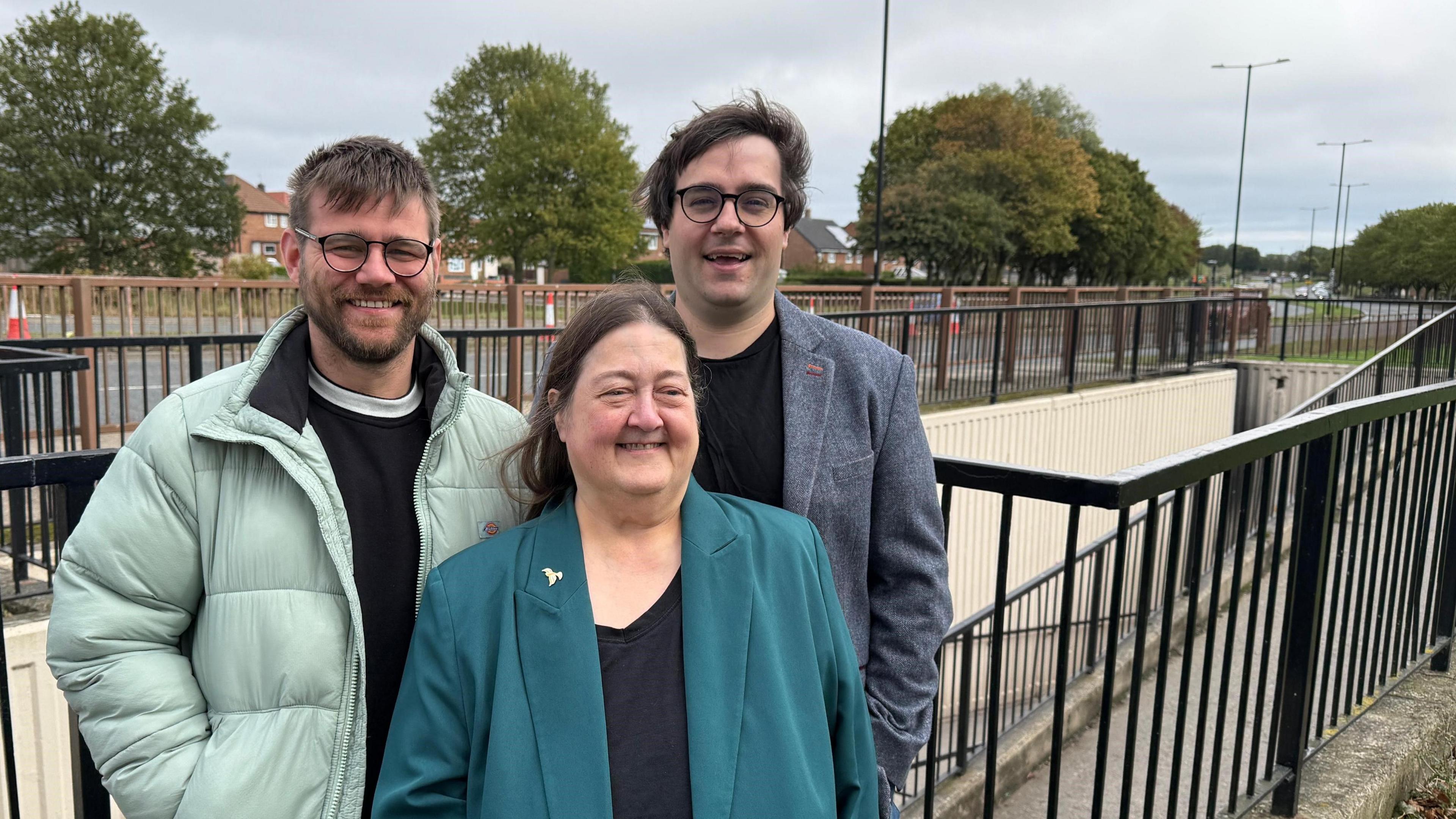 Liberal Democrat councillors Stephen O’Brien, Margaret Crosby and Paul Edgeworth smiling at the camera in front of an underpass. O'Brien is wearing a green puffer jacket and a black jumper. He has round glasses and blonde hair and beard. Crosby is wearing a teal suit jacket and a black top. She has long brown hair parted in the middle. Edgeworth is wearing a grey tweed jacket and a black top. He has round glasses and brown hair swept to the side.