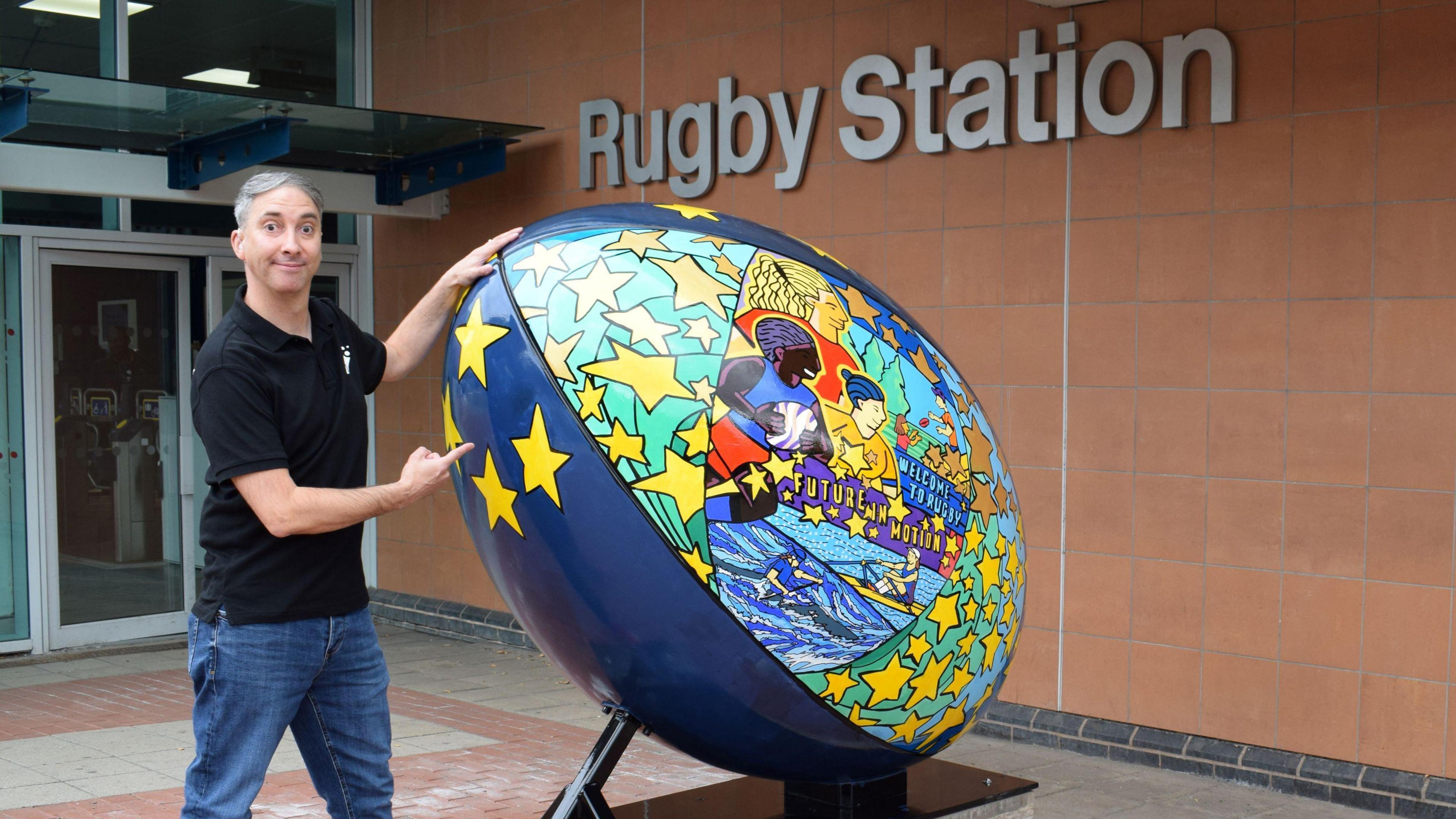 Olivier Jamin wearing a black t-shirt and jeans standing next to a 6ft colourfully decorated rugby ball next to a Rugby Station sign