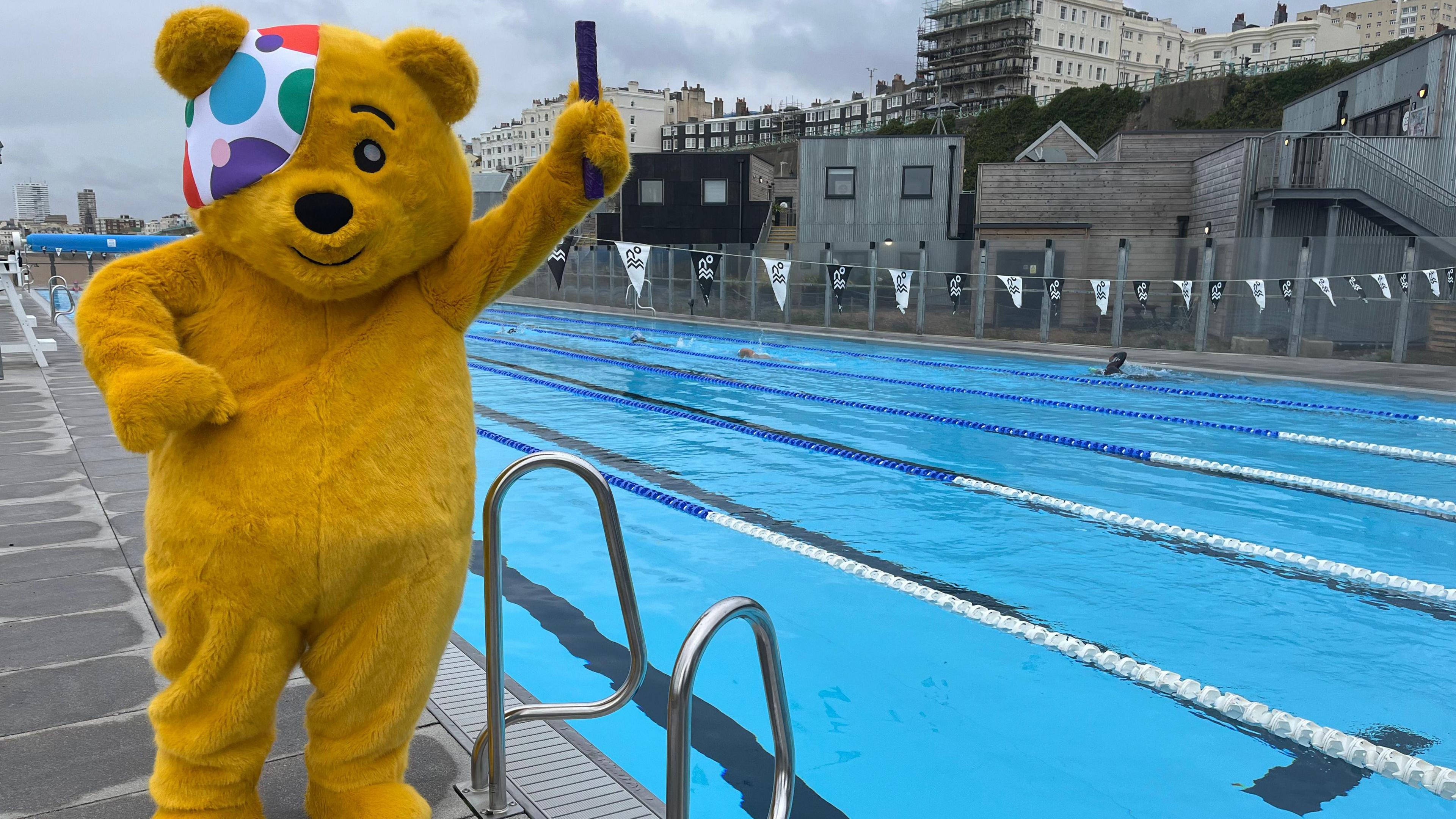 Pudsey, the Children in Need mascot, holding a relay baton standing next to a swimming pool