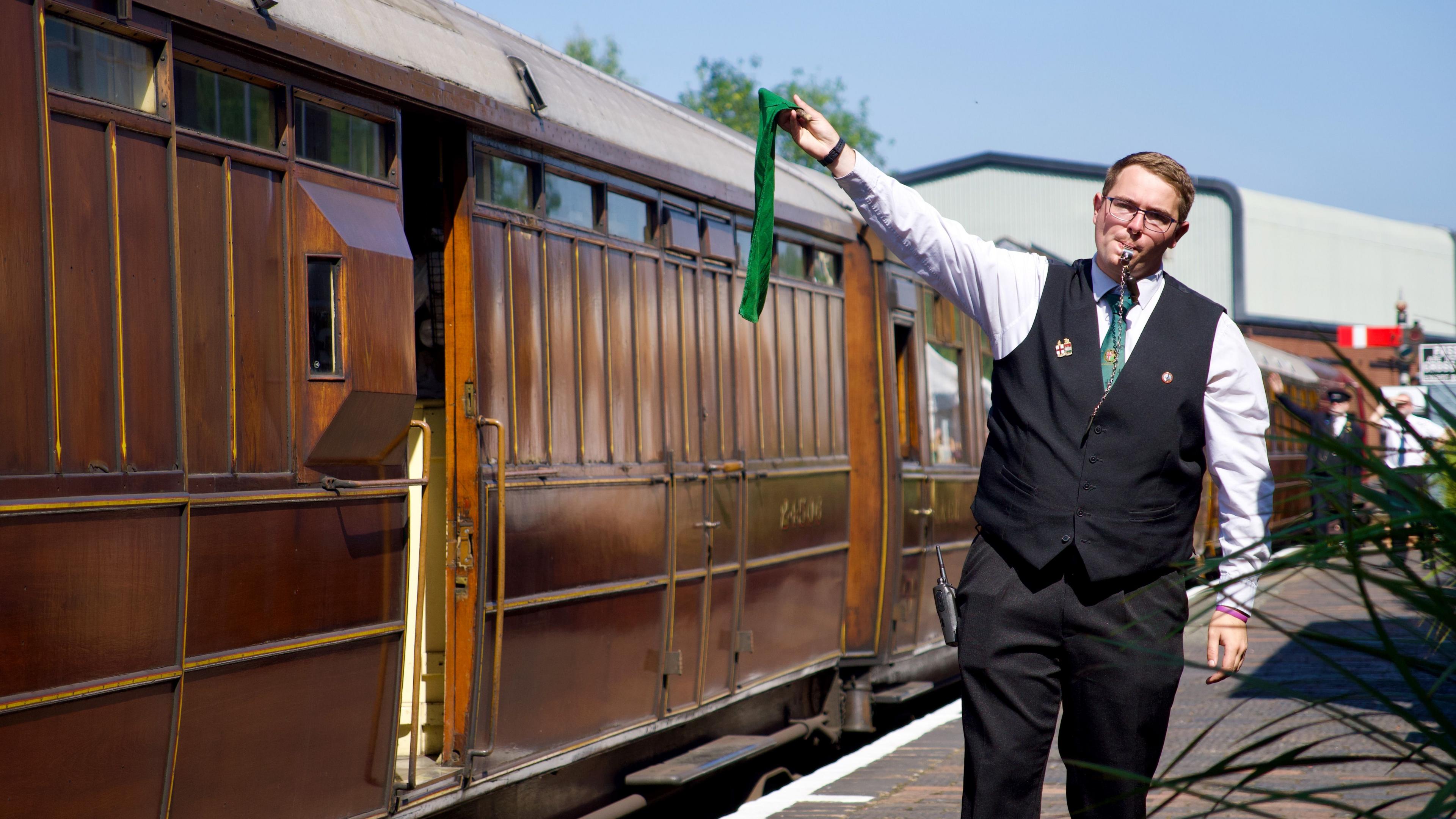 A railway guard, with a whistle in his mouth, waves a green flag to signal the departure of Flying Scotsman. The guard, who has glasses, is wearing black trousers, black waistcoat and white shirt with green tie