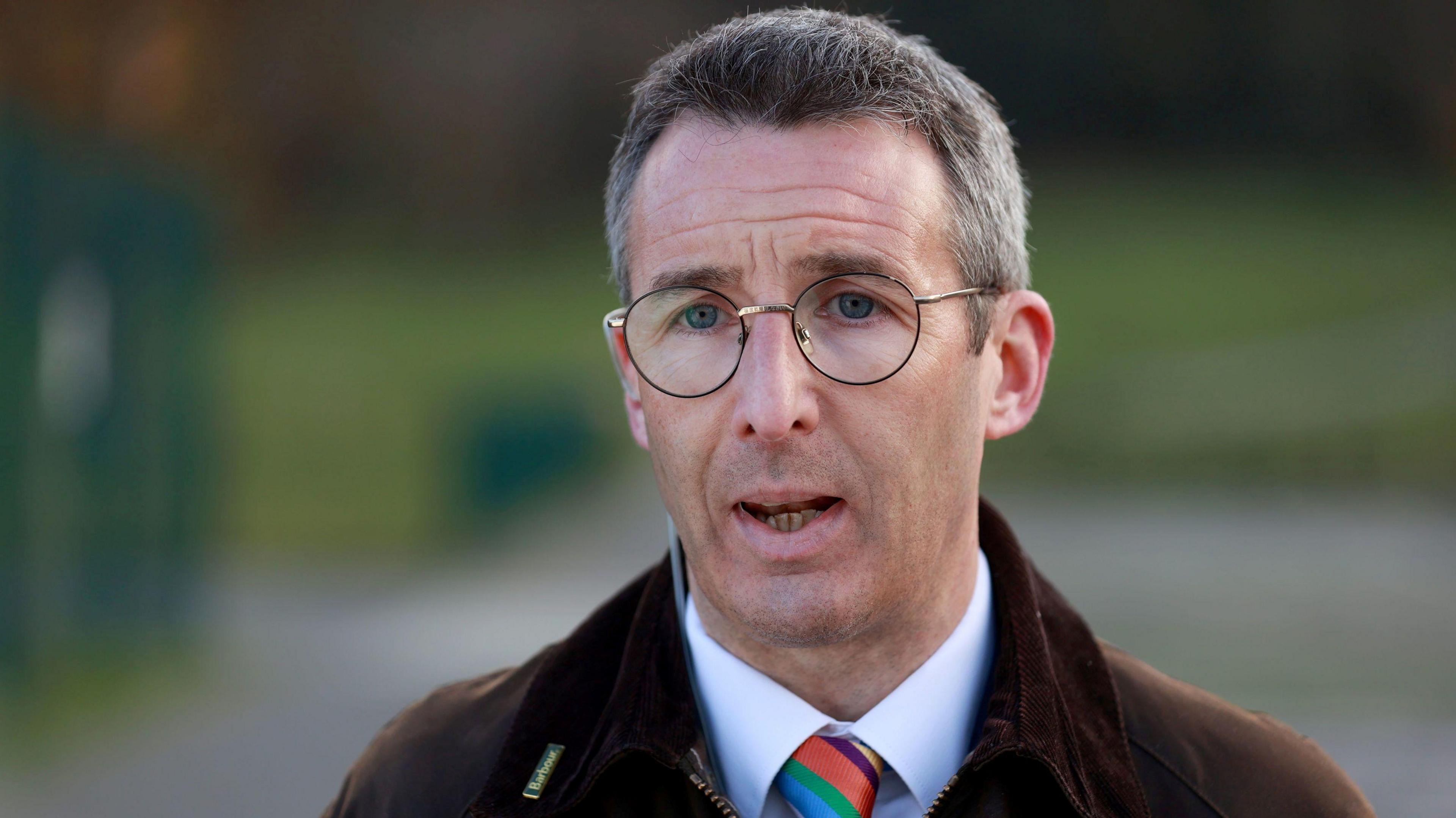 Muir is talking to the media. The background is blurred. He wears round glasses and has a brown jacket on with a colourful tie and a white shirt.
