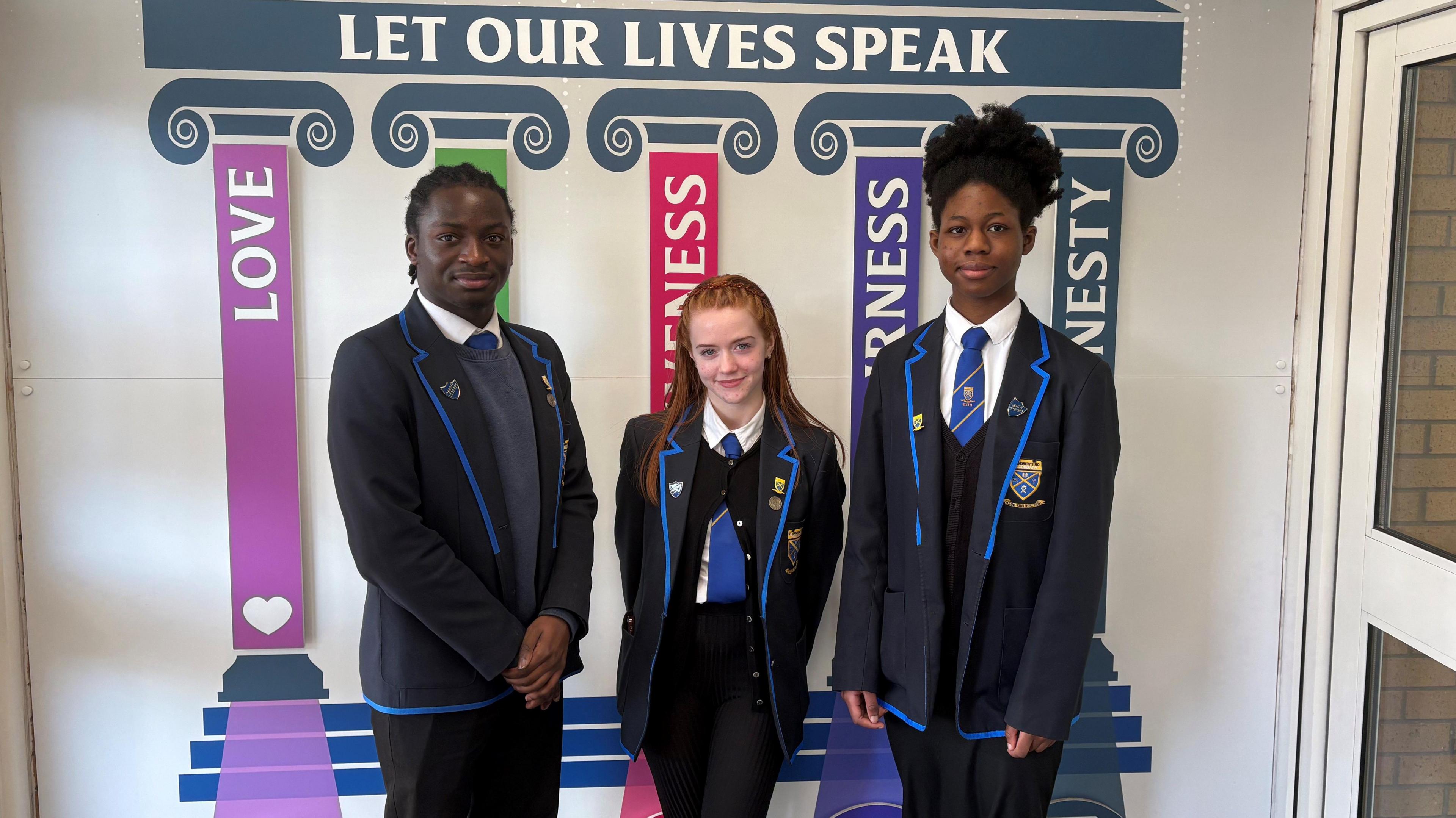 Pupil David, Lucy and Ayomide in school uniform standing in front of pillars painted on a white wall with writing on it.