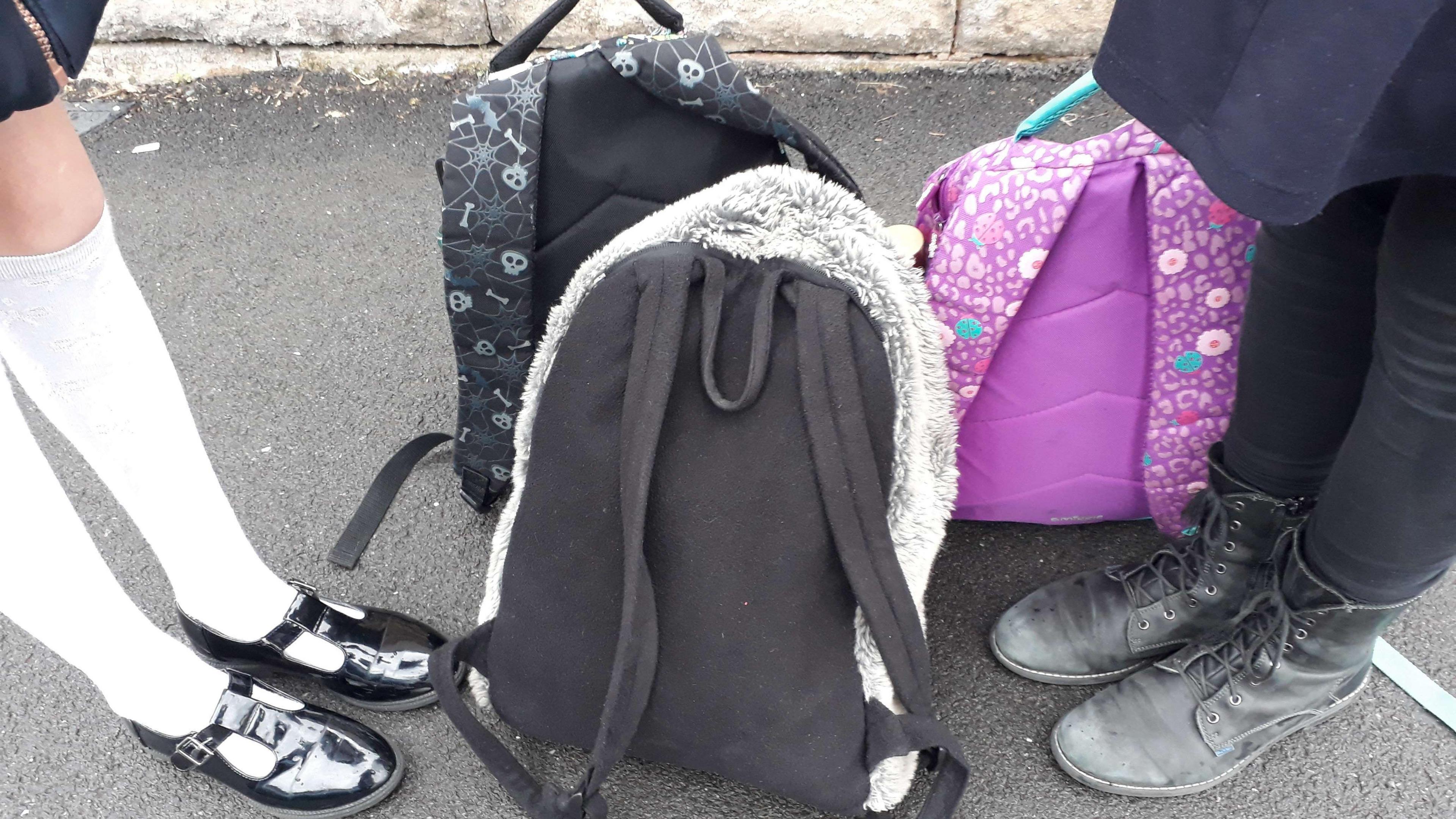 Two sets of feet - one dressed in white socks and black patent shoes, the other in black leggings and black lace-up boots, are standing on the concrete. Three rucksacks, in various colours, are on the floor between their feet. 