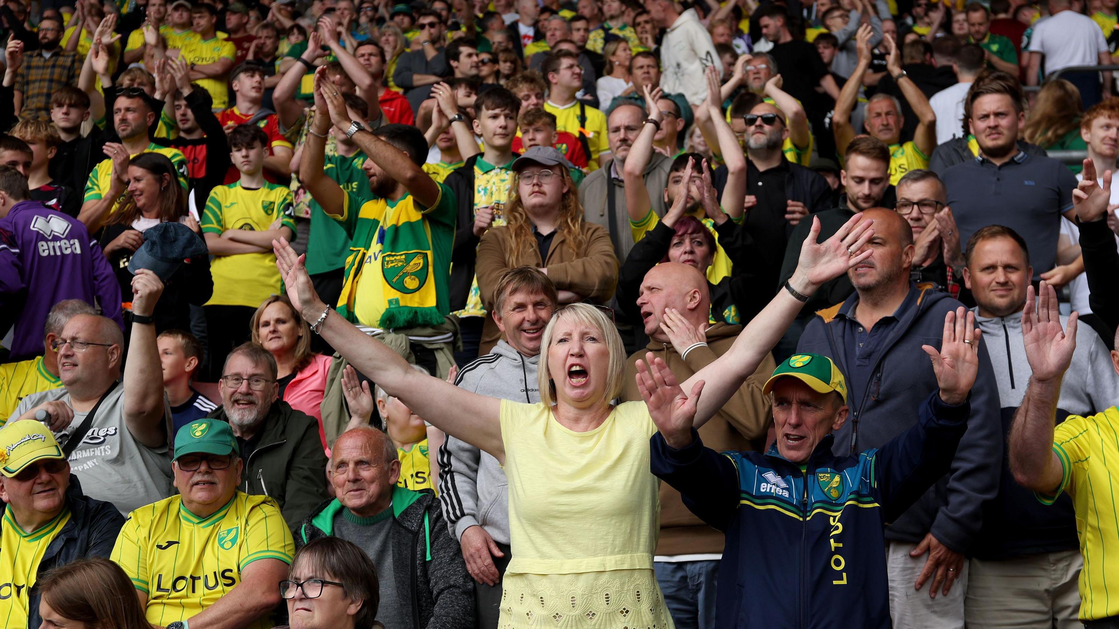 Dozens of football fans are shown in a stadium stand, with many wearing the yellow shirt of their team and some standing with their hands aloft as they sing.