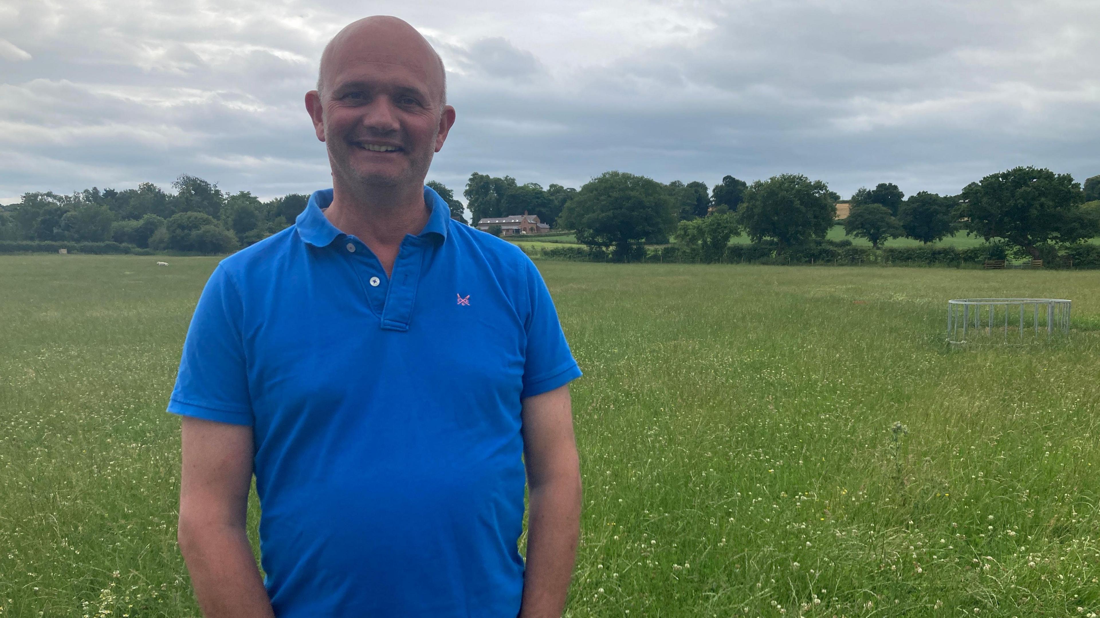 Farmer Phil Latham is standing in a field at his farm in Chorley, near Nantwich. He is bald and is wearing a blue polo shirt.