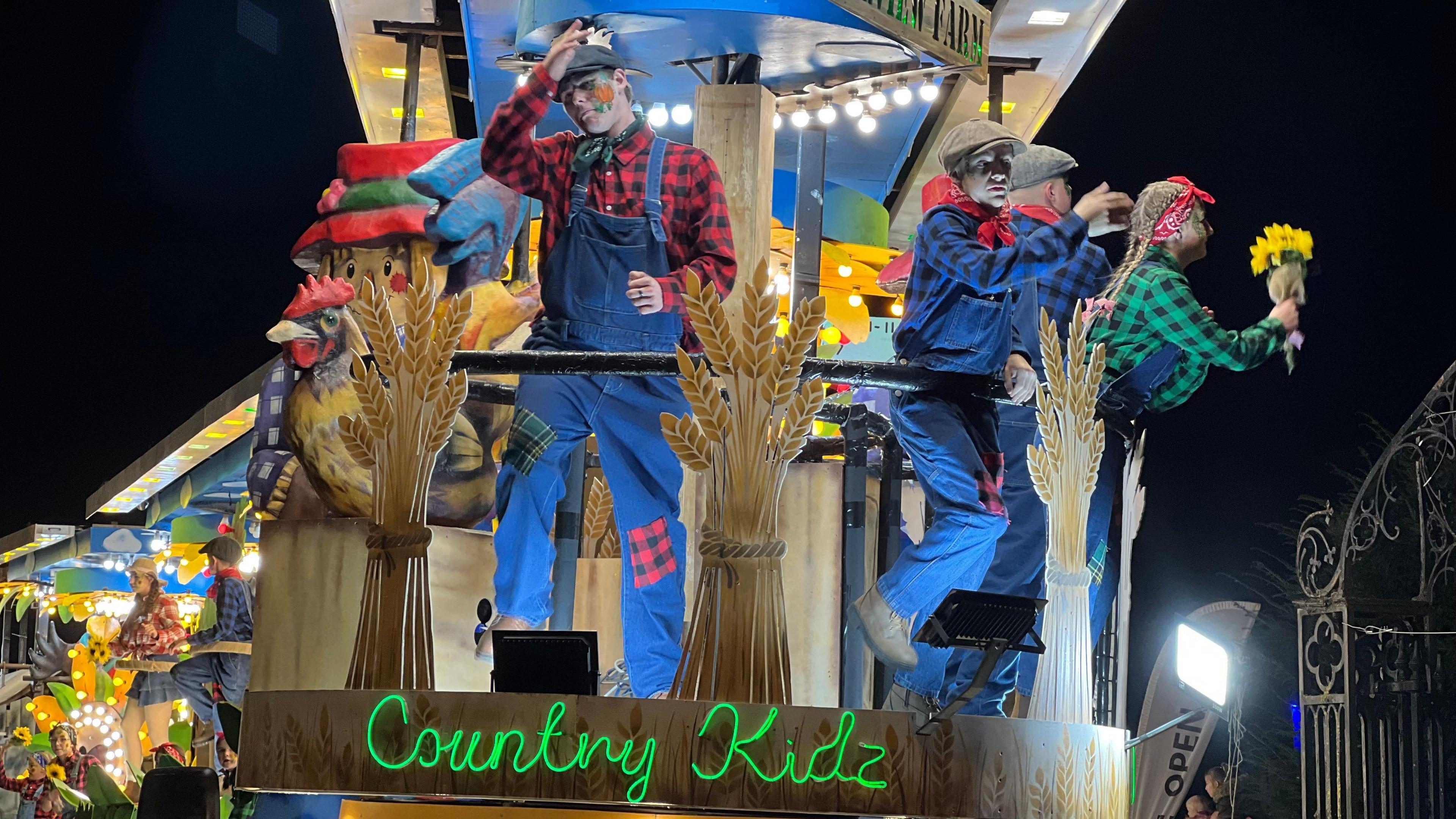 People dressed as scarecrows stand on top of a cart, which has a green neon sign that says "country kidz" attached to the front of it