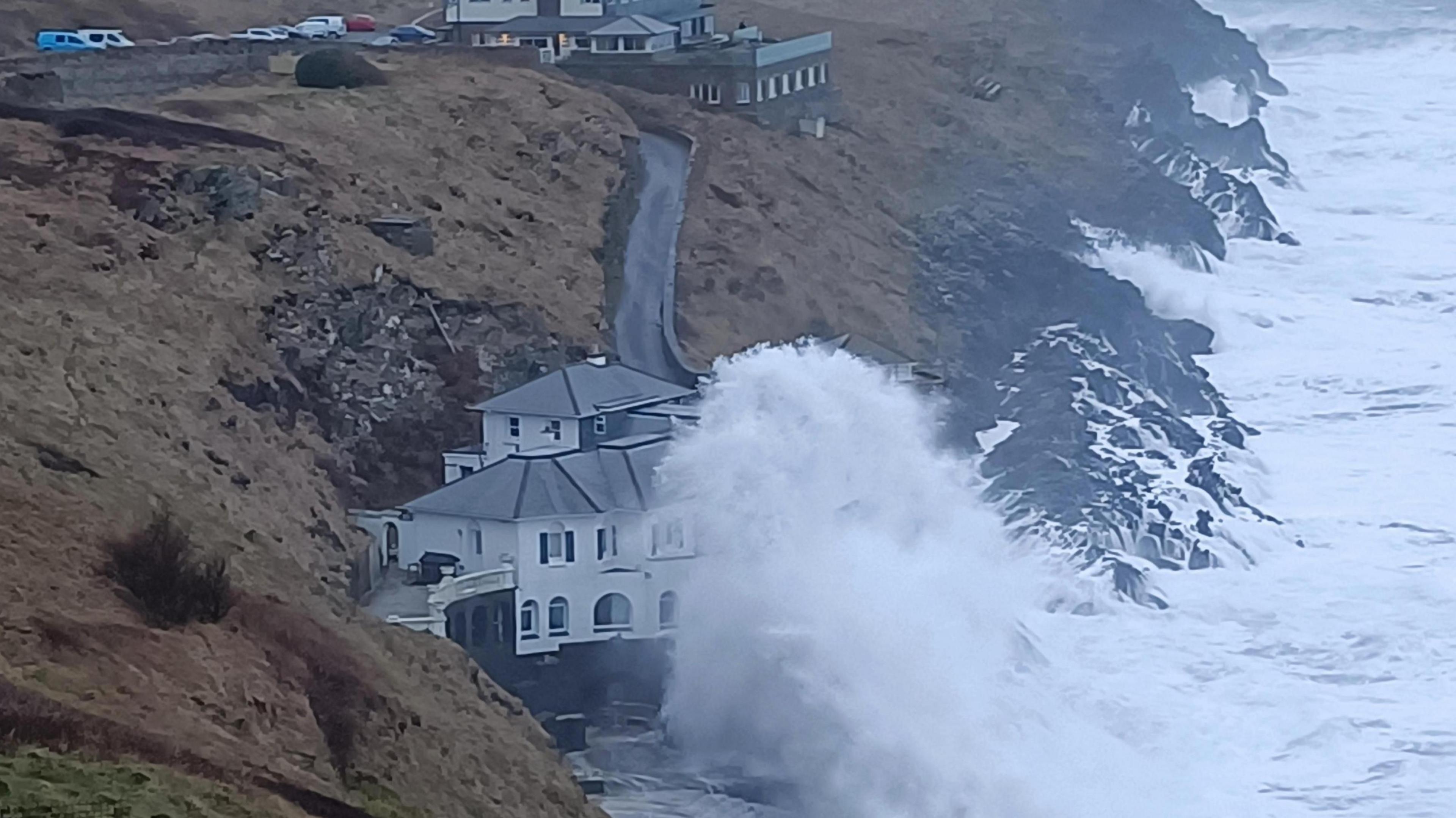 Big waves crash against a white house at the bottom of a coastal path on cliffs next to the sea