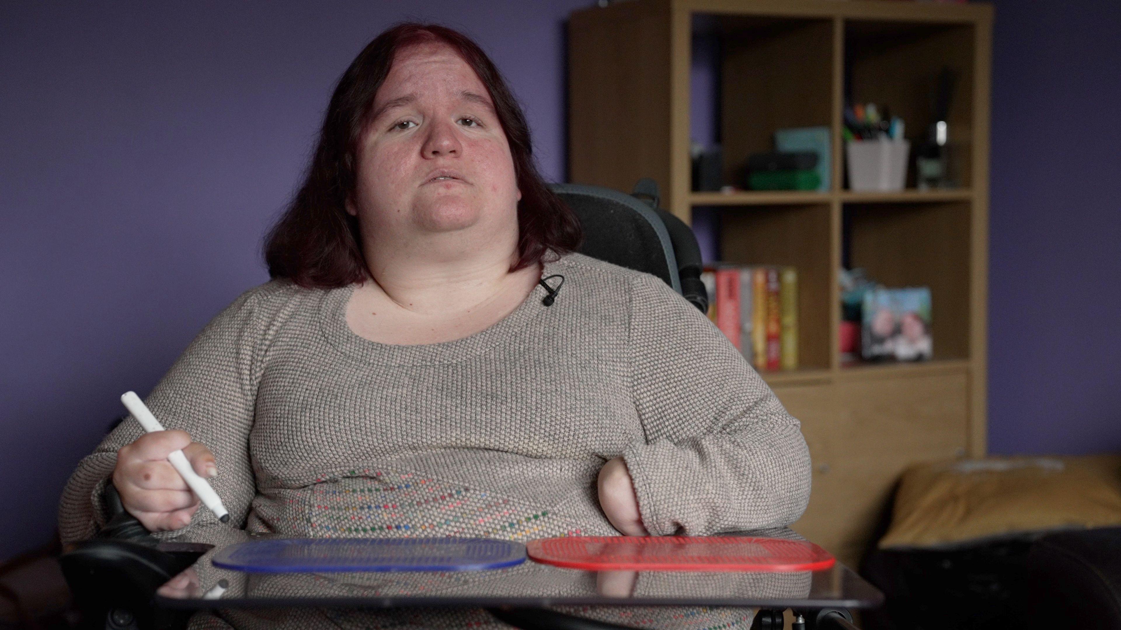 Emma in her living room which is decorated purple. Emma is sitting on her electric wheelchair looking into the camera