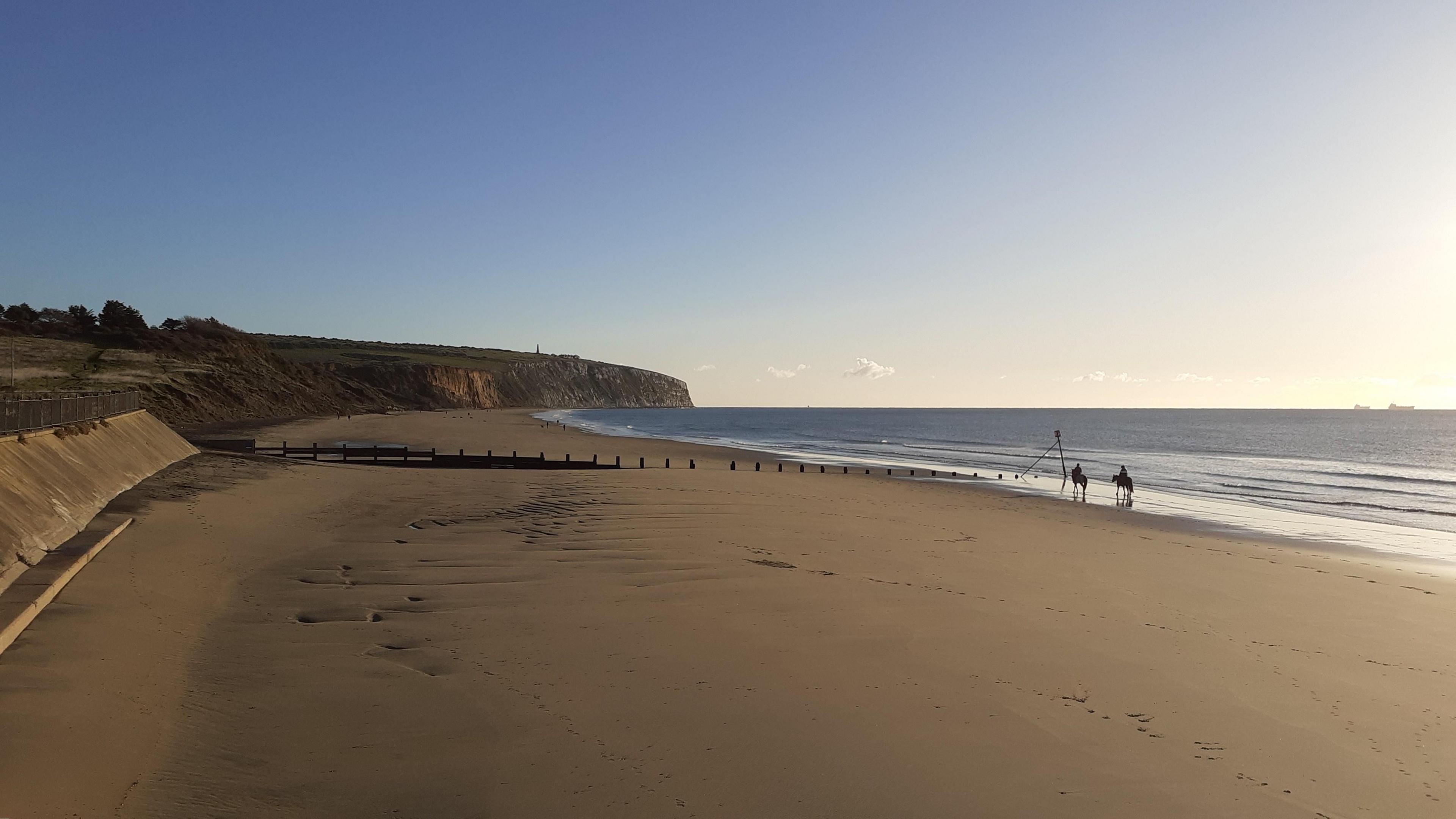 A wide open view of a beach with golden sand washed clean by the sea which has now receded. A wooden groyne can be seen half way down the beach with the bending coastline behind it. The sky is bright blue and cloud free. Two horses with riders can be seen walking through the water's edge and two shipping tankers are on the horizon.