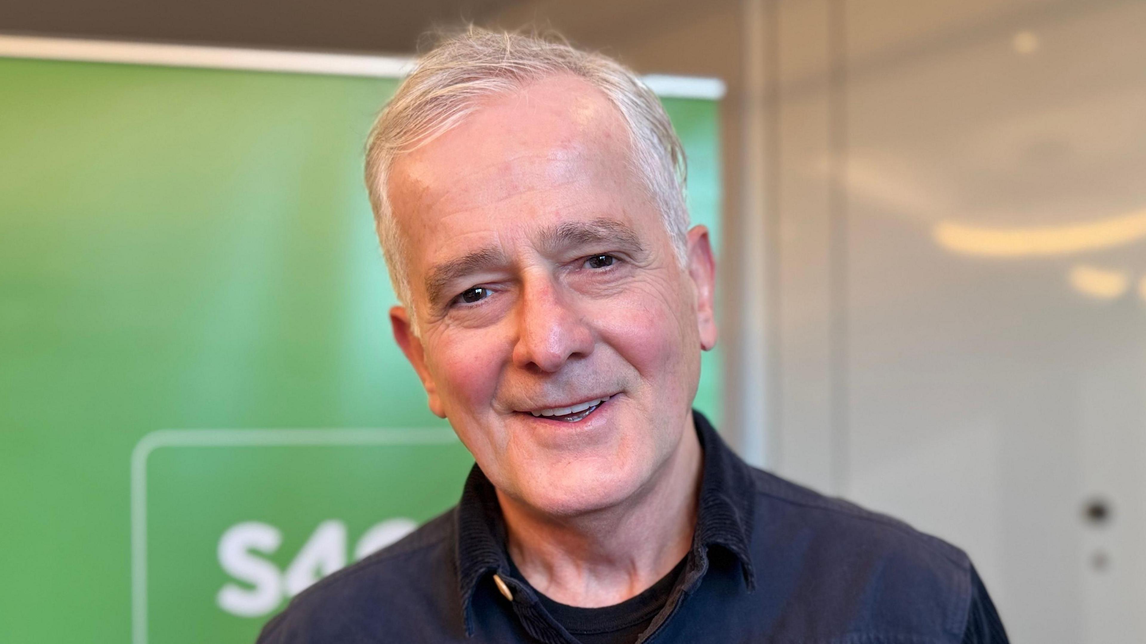 Huw Chiswell sydd yn y llun yma. Mae e'n gwenu wrth edrych ar y camera ac yn gwisgo crys glas tywyll. Mae ganddo wallt gwyn.