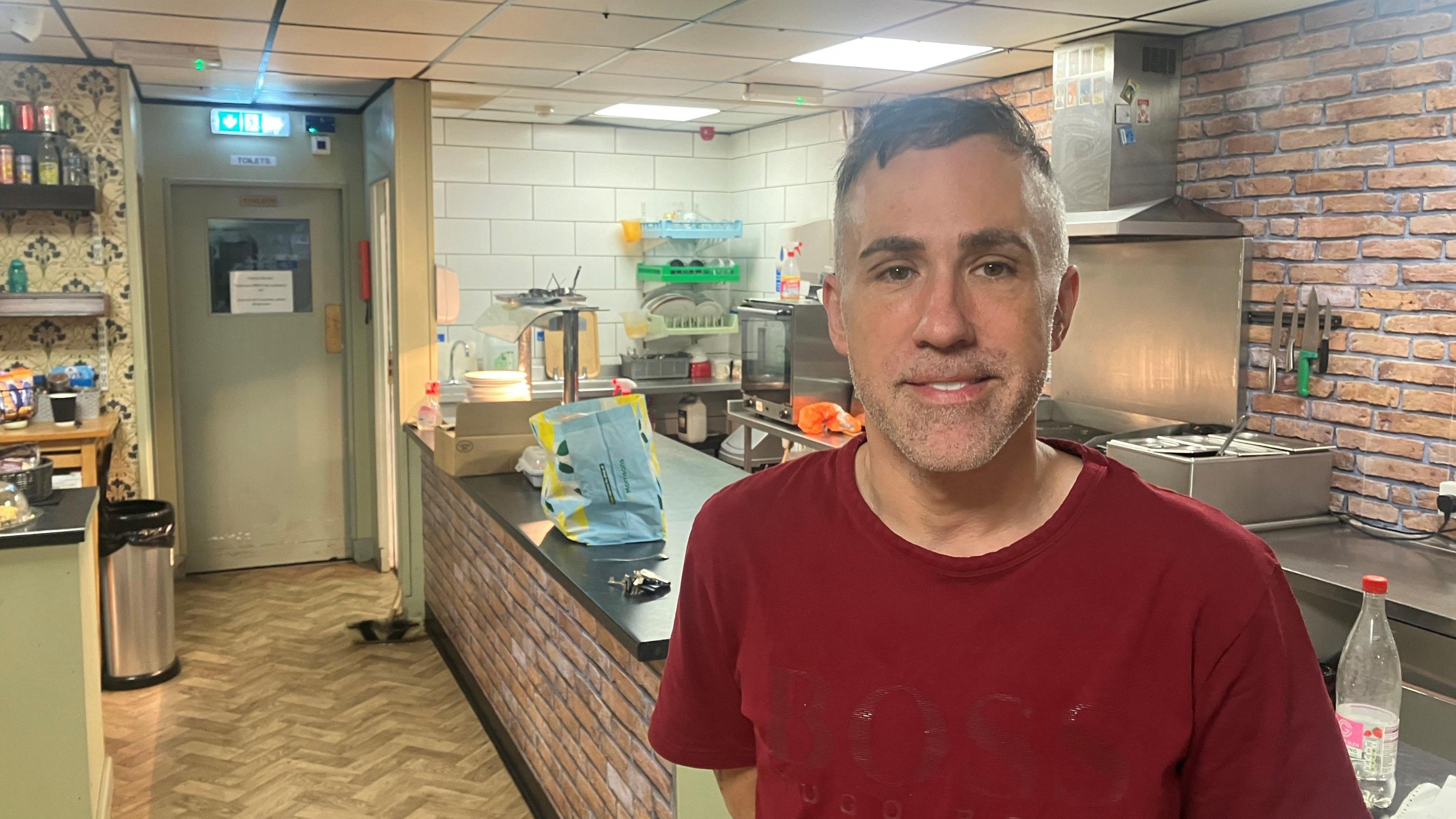 Andy Flaherty in the kitchen of his community cafe. He is wearing a red t-shirt and has short, cropped dark hair and stubble. He stands in front of a counter with a hob, grill and hot plate in the background.