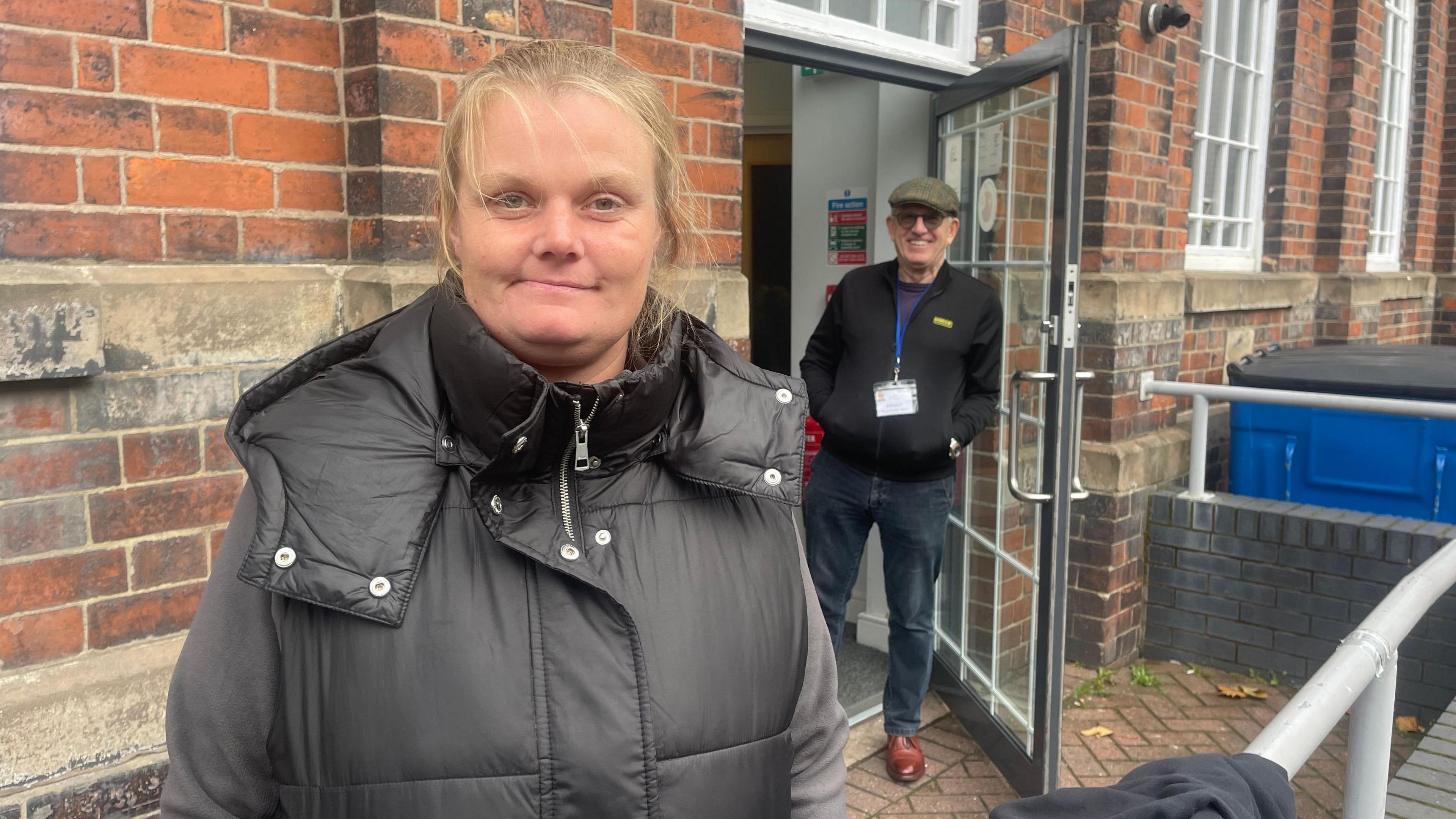 A woman is stood outside a brick building. The door is being held open by a man who is smiling. The woman has fair hair tied back, and is wearing a black body warmer and a grey jumper. The man has on a flat cap, sunglasses, black jacket, dark jeans and red shoes.