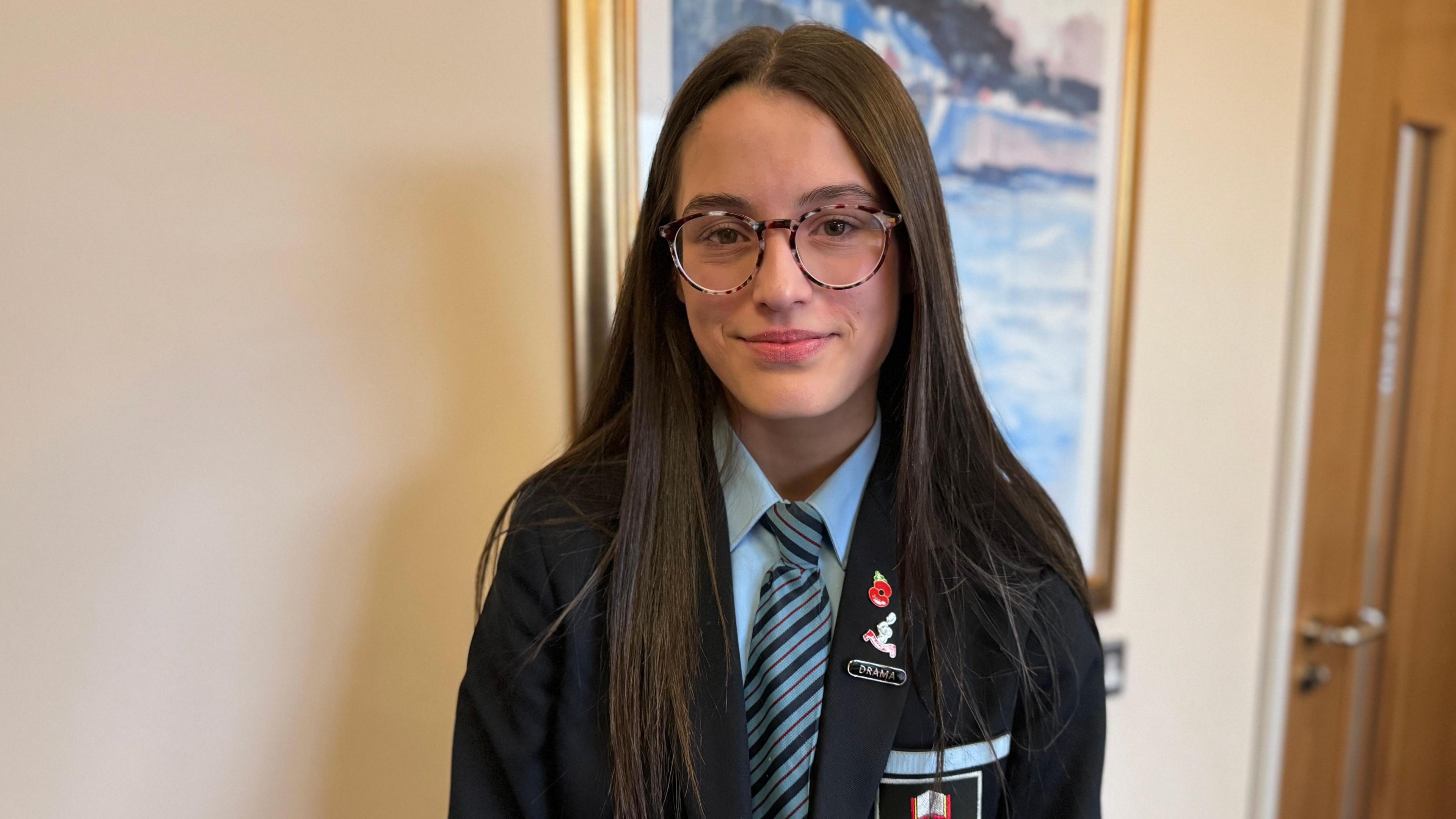 Pippa smiles in her school uniform. She has long brown hair and glasses. On her navy blazer there is a red poppy pin.