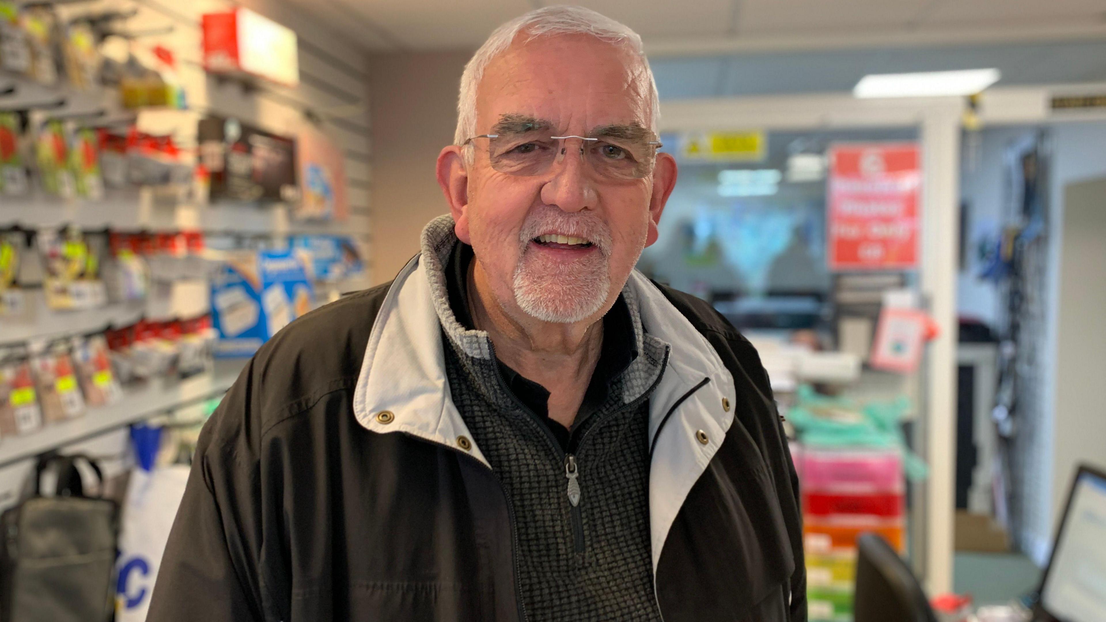 A man with grey hair and glasses, wearing a black winter jacket and green sweater, standing in a stationary shop. 