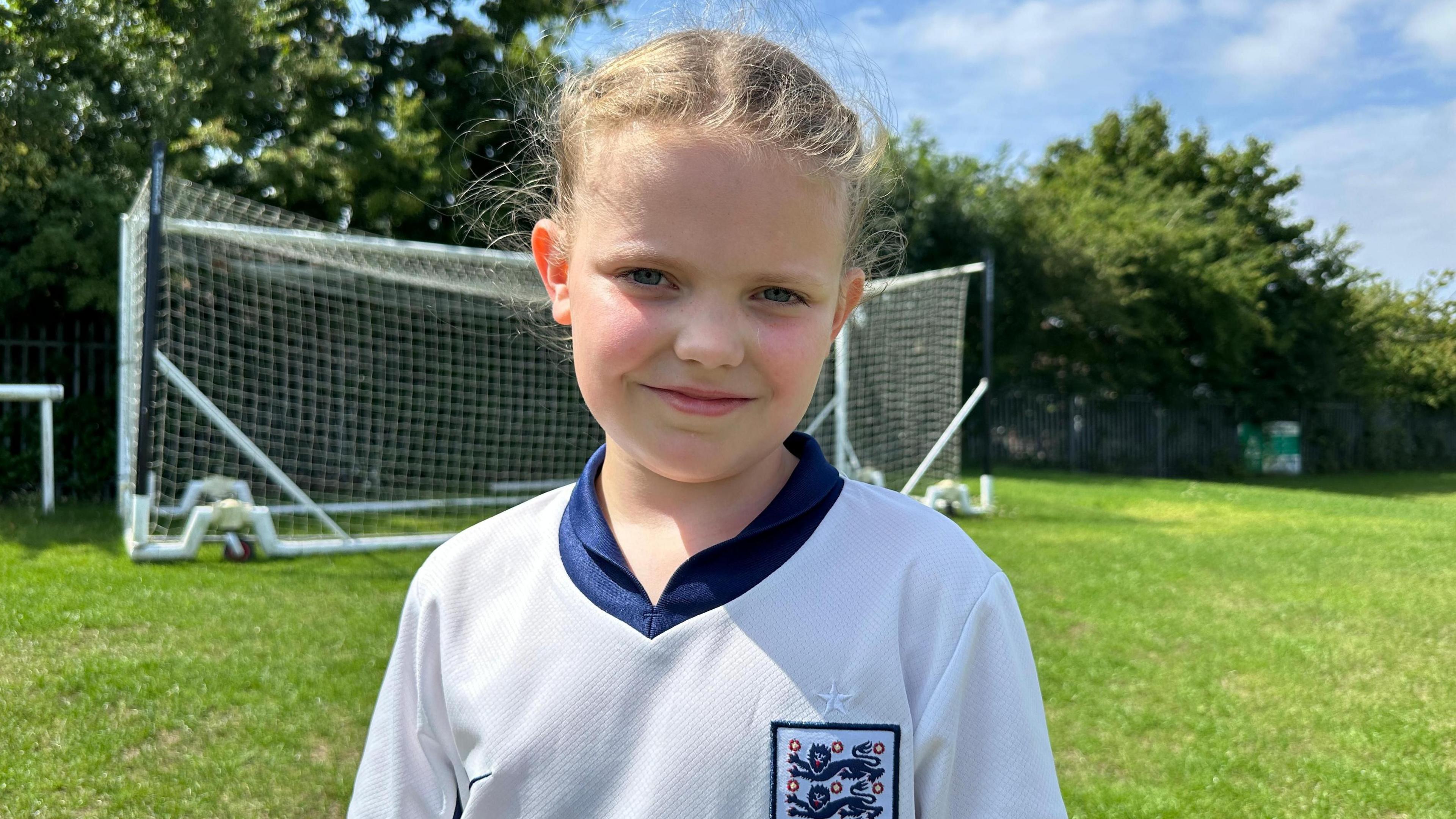 Girl with blonde hair is standing on a football pitch with a net behind her and is wearing a white England football shirt with a three Lions England badge and blue collar.