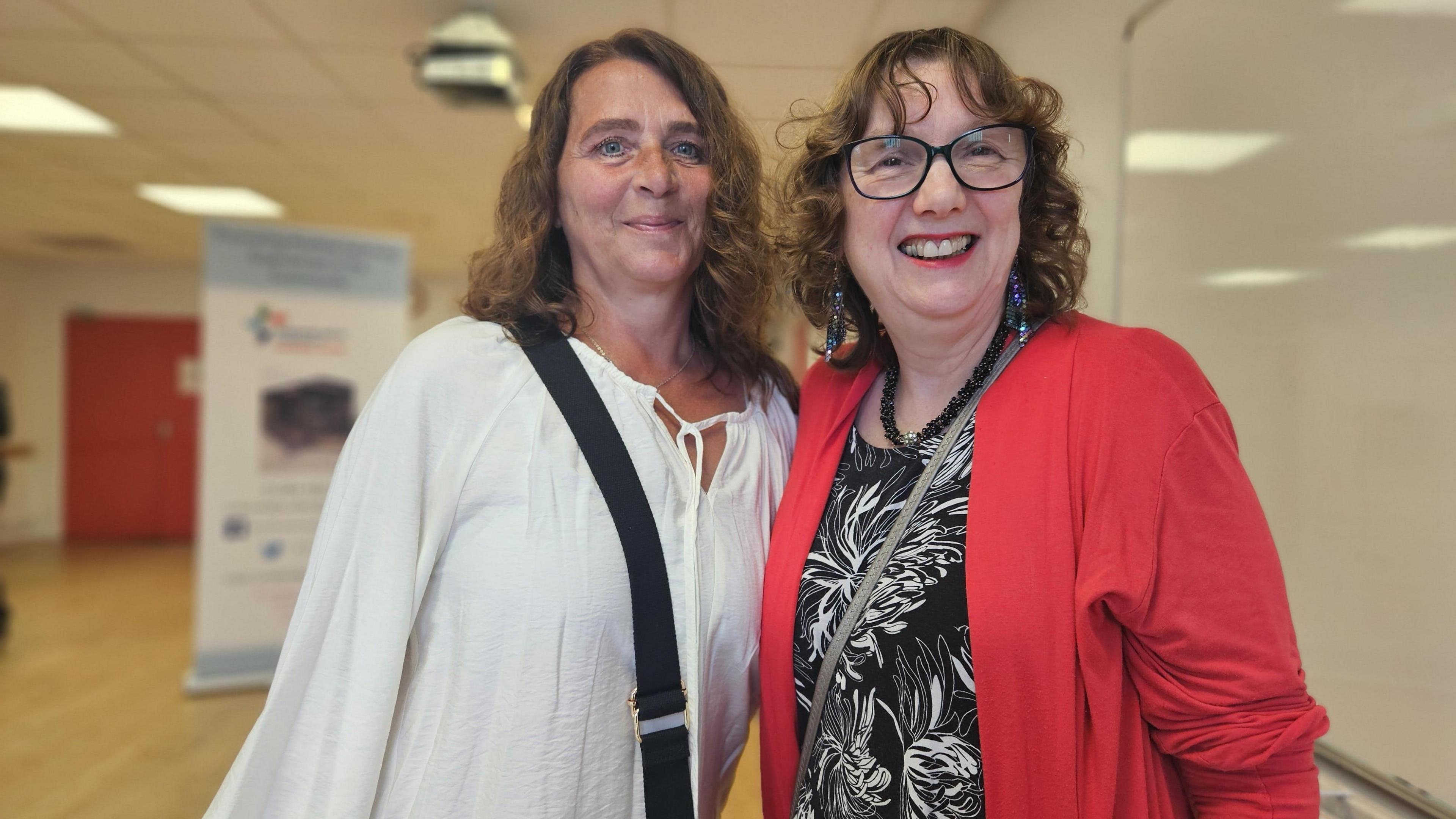 Two middle-aged women smile into the camera. They are standing in a community hall. Both have short brown/dark blonde hair. One wears a white cotton shirt, the other wears a red jacket, thick glasses and big earrings.