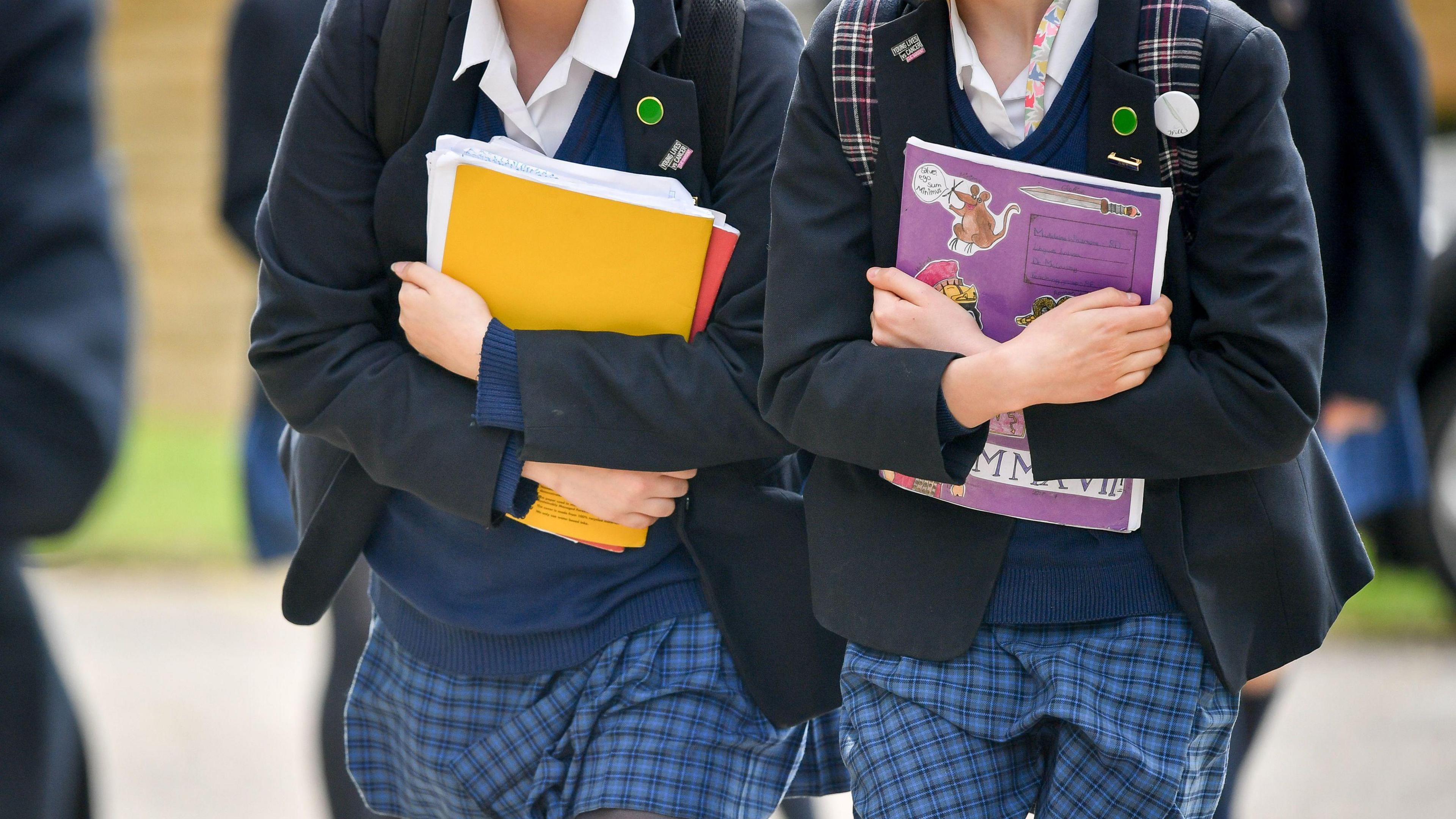 Photo of two school girls walking to school in school uniform. Both girls are carrying text books with rucksacks on their backs. 