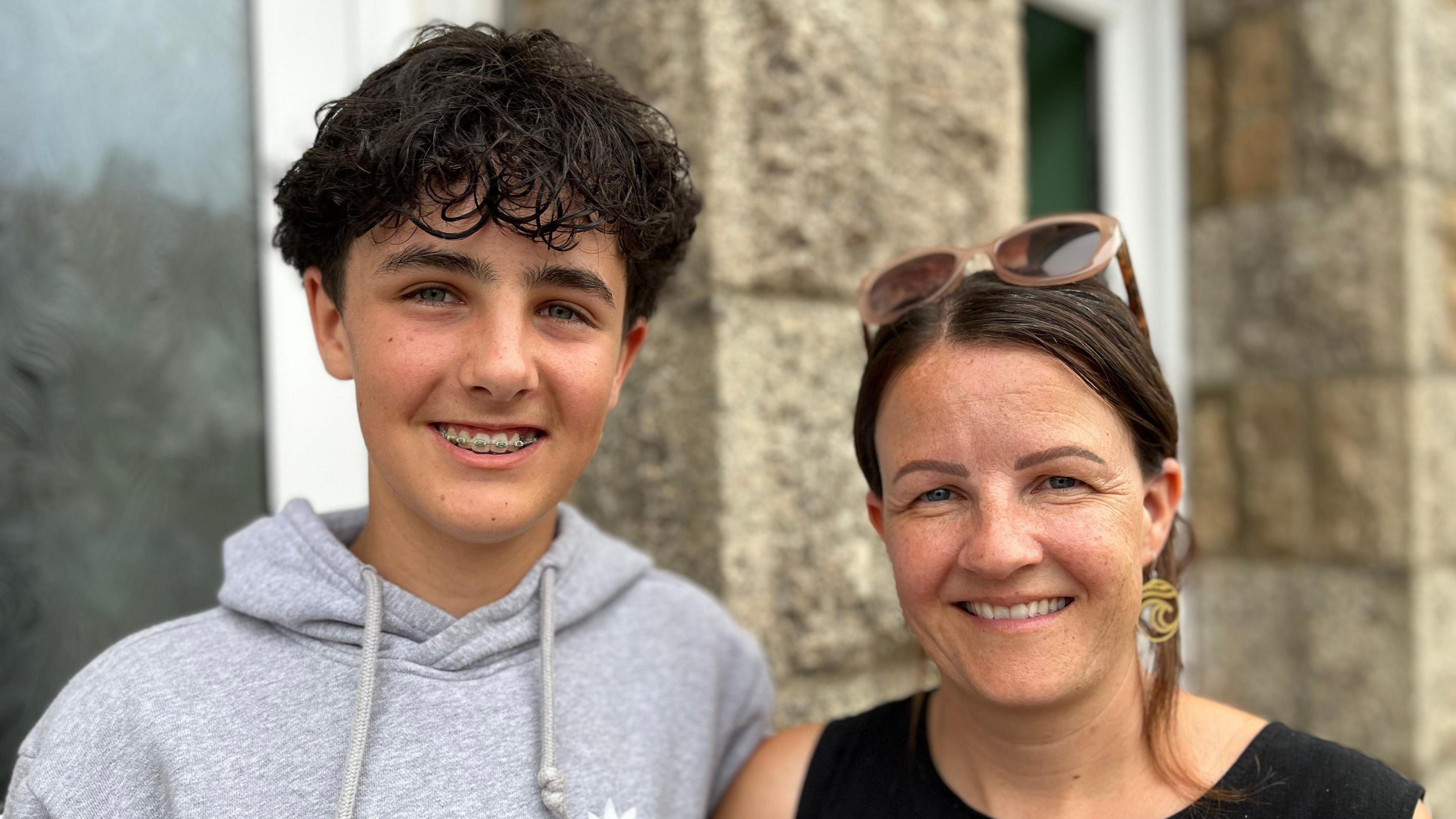 A close-up portrait photo of Jacob and his mother Amy standing shoulder to shoulder outside a stone building; one has dark curly hair, wears a grey hoodie and smiles with braces visible, while the other has dark hair tied back, sunglasses resting on their head and gold earrings.

Both are looking directly at the camera, with their faces in sharp focus against a softly blurred background of stone walls and large windows.