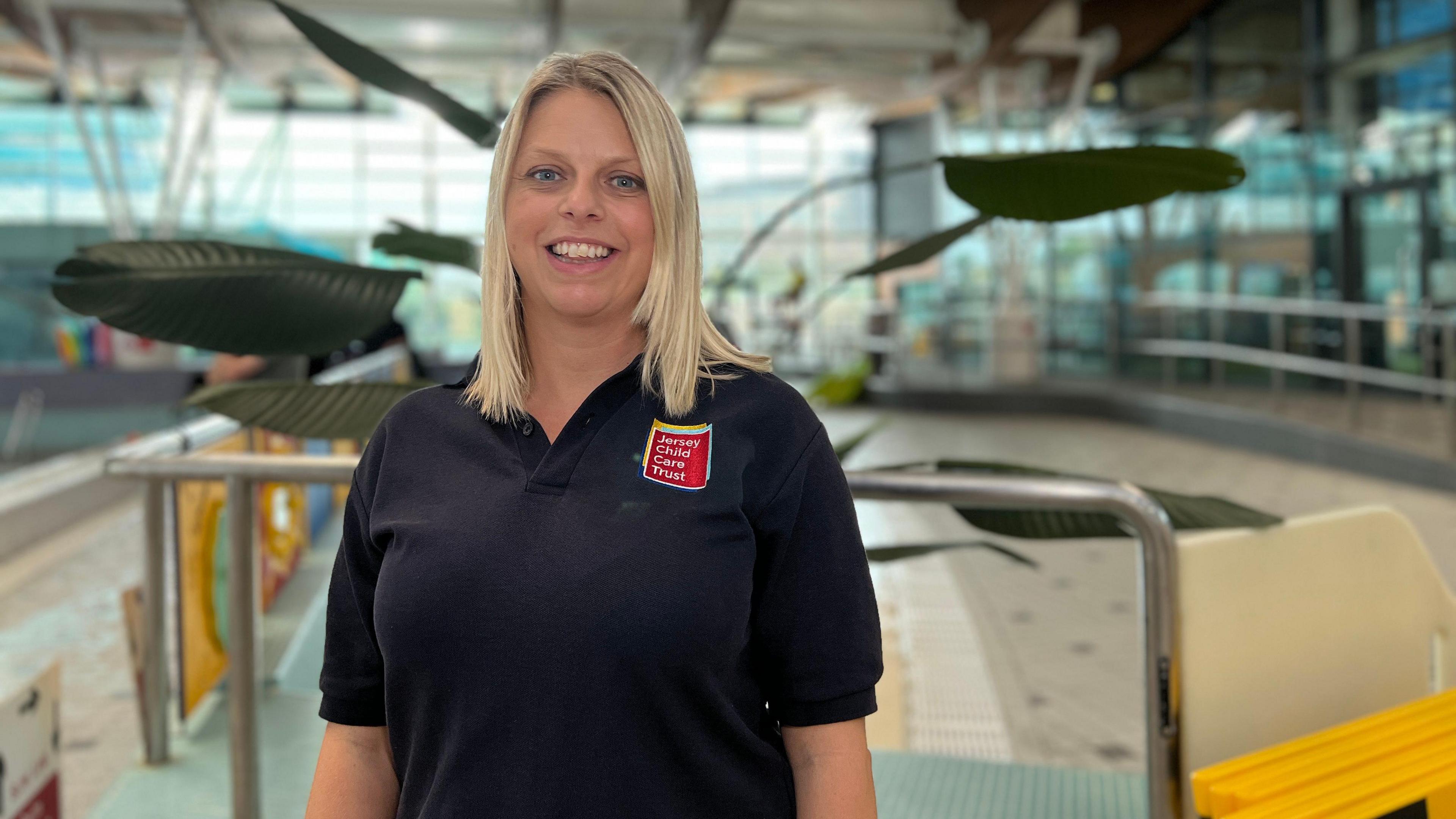 Alison smiles at the camera as she stands in the pool side area. She's wearing a navy blue top with a red logo of the Jersey Child Care Trust on her chest. She has shoulder length blonde hair. Behind her is a big plant with large leaves.