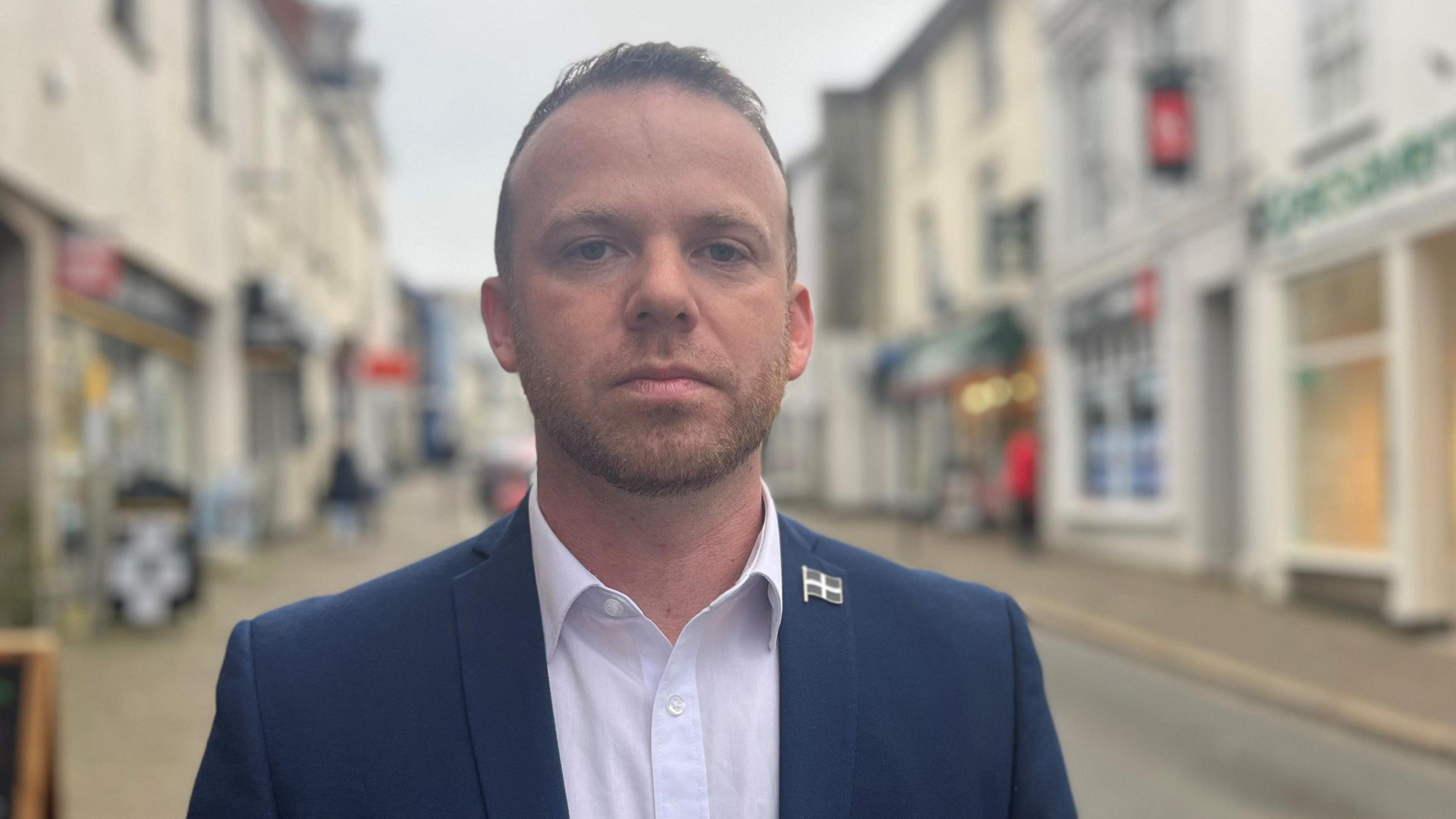 A man with a short beard standing in a shopping street looking straight at the camera wearing a blue jacket, white shirt and Cornish flag pin badge 