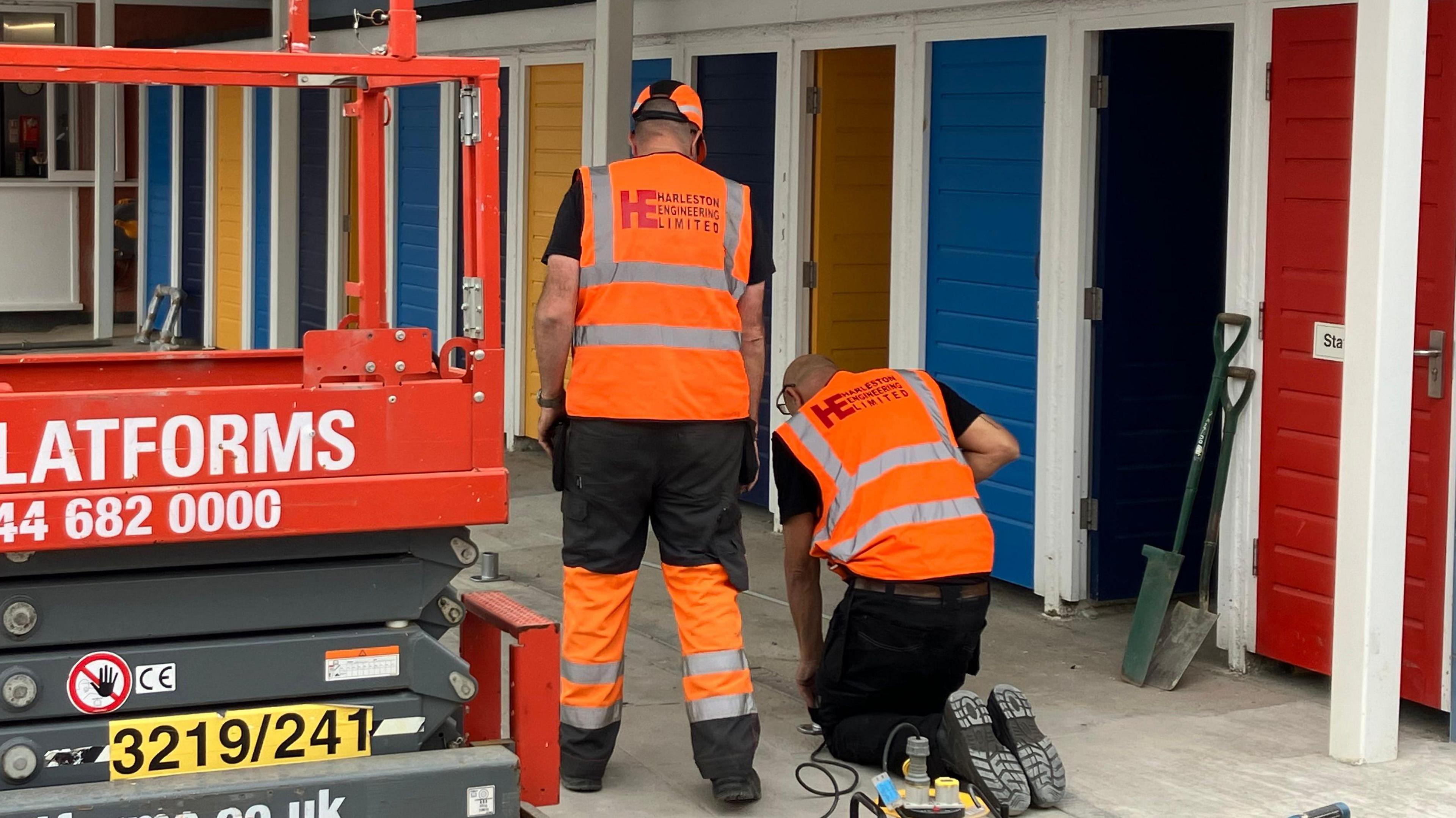 Two workers, one standing and one kneeling, are wearing orange fluorescent clothing and working on the base of the new canopy at the pool, surrounded on one side by brightly-coloured changing room doors 