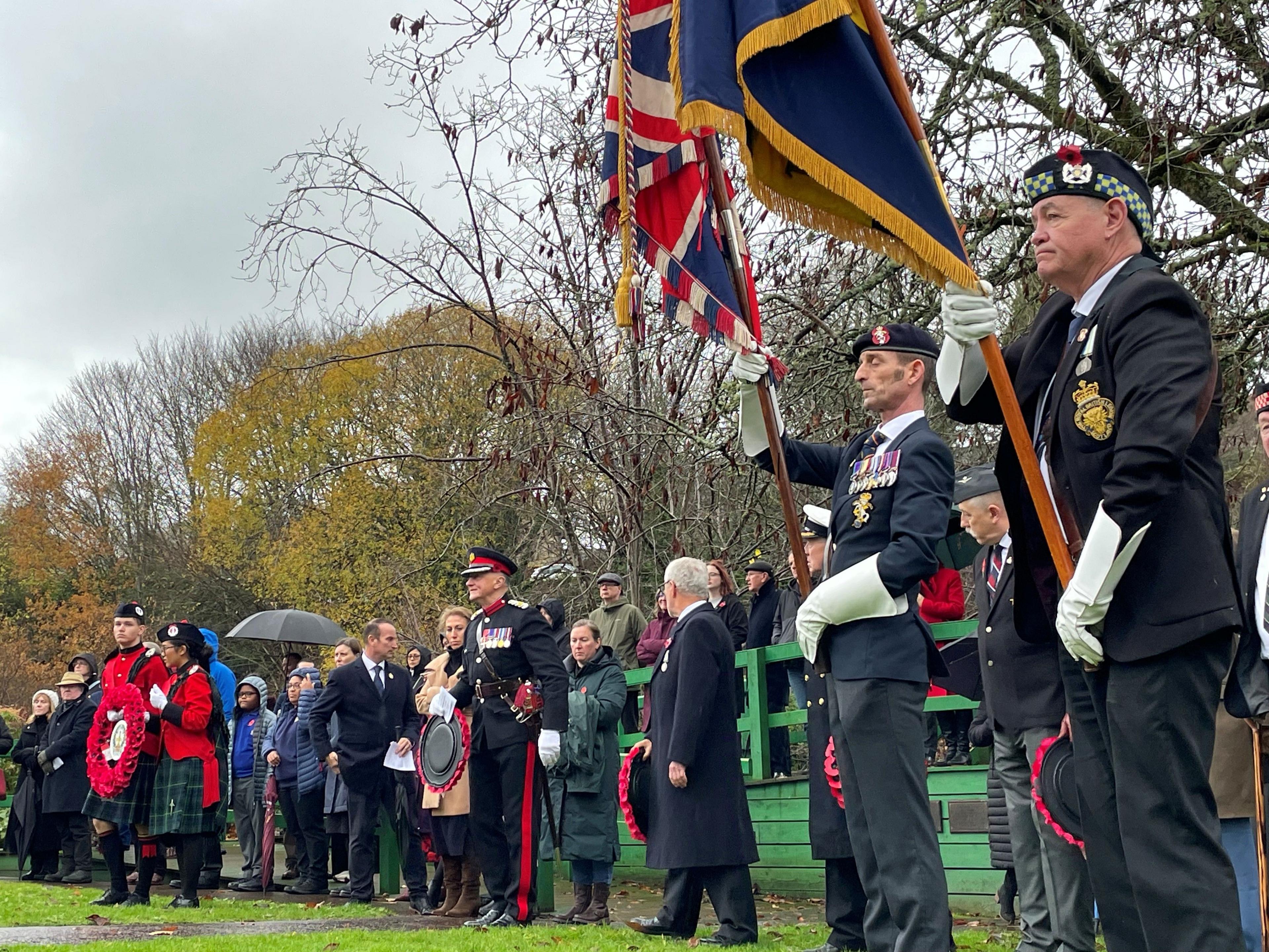 People, some in military uniform and carrying banners, pay their respects, with trees in the background