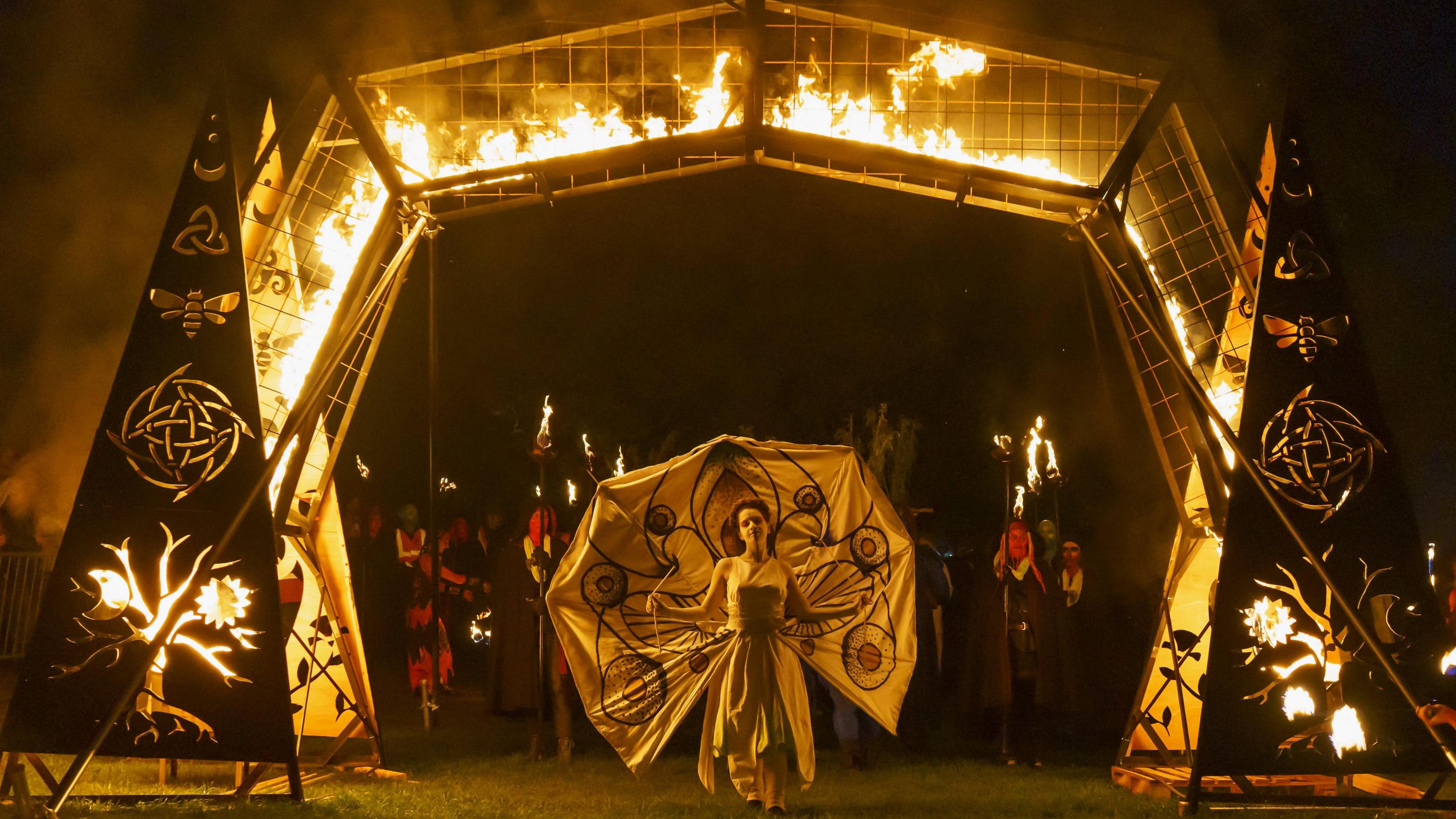 The May Queen wearing a white dress walks through a gate surrounded by fire at the start of the Beltane festival.