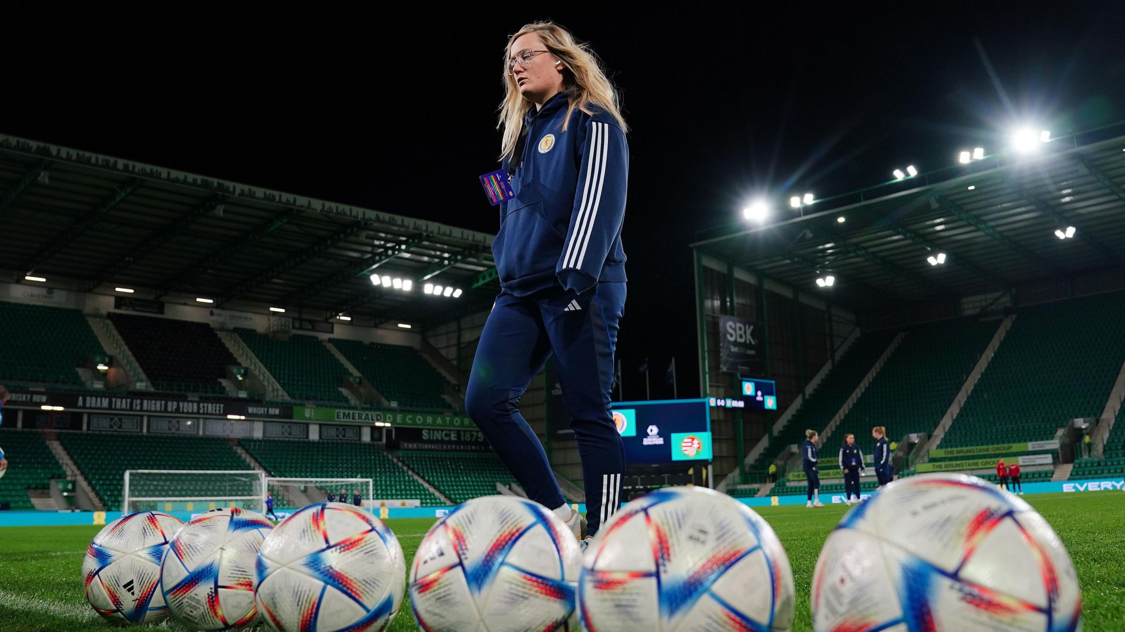 The image shows Scotland player Erin Cuthbert in training kit stood in an empty stadium in front of a line of footballs. 