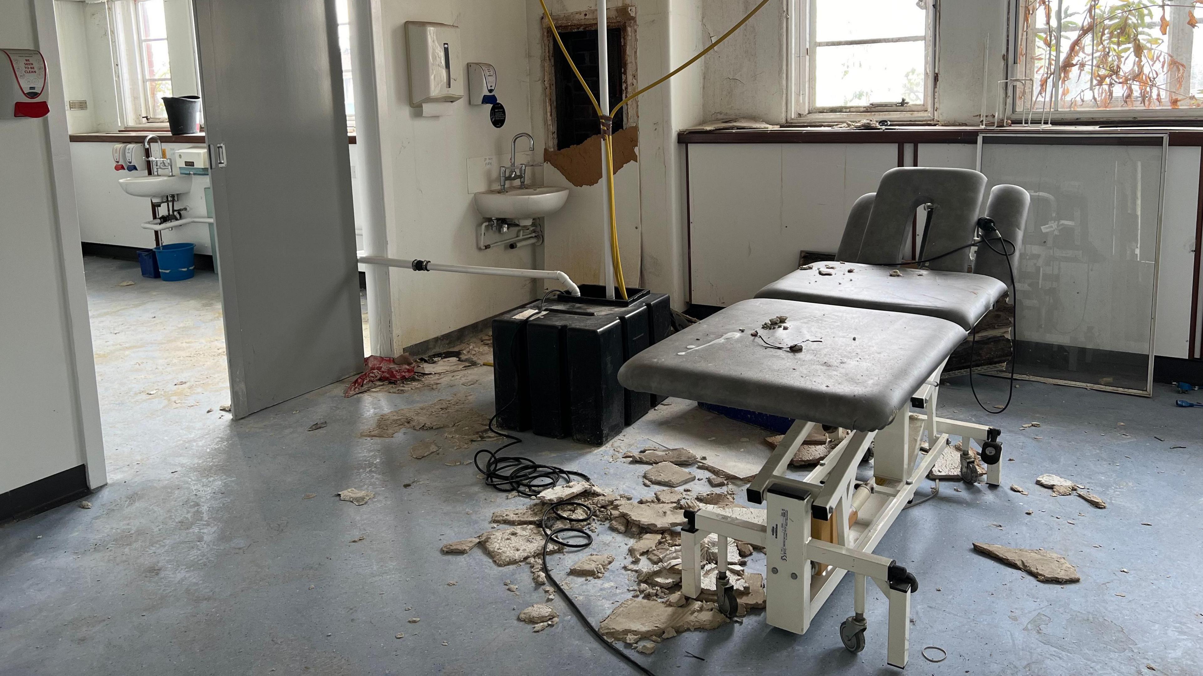 A disused hospital bed surrounded by debris, a broken wall and a muddy floor. 