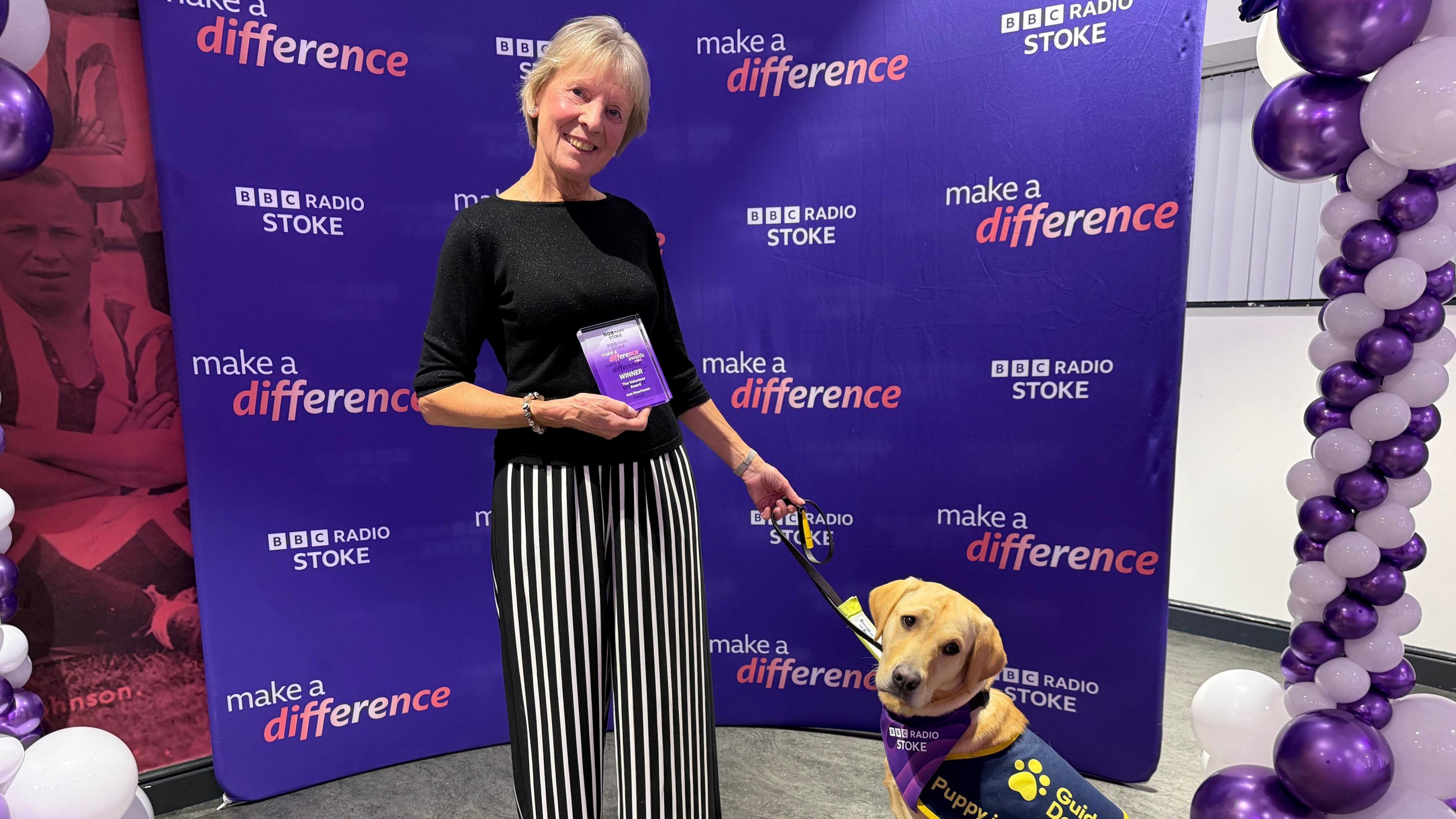 Ann Moorhouse, a woman wearing a black top and back and white striped trousers with guide dog. They are positioned in front of a purple BBC Radio Stoke Make A Difference step and repeat board.