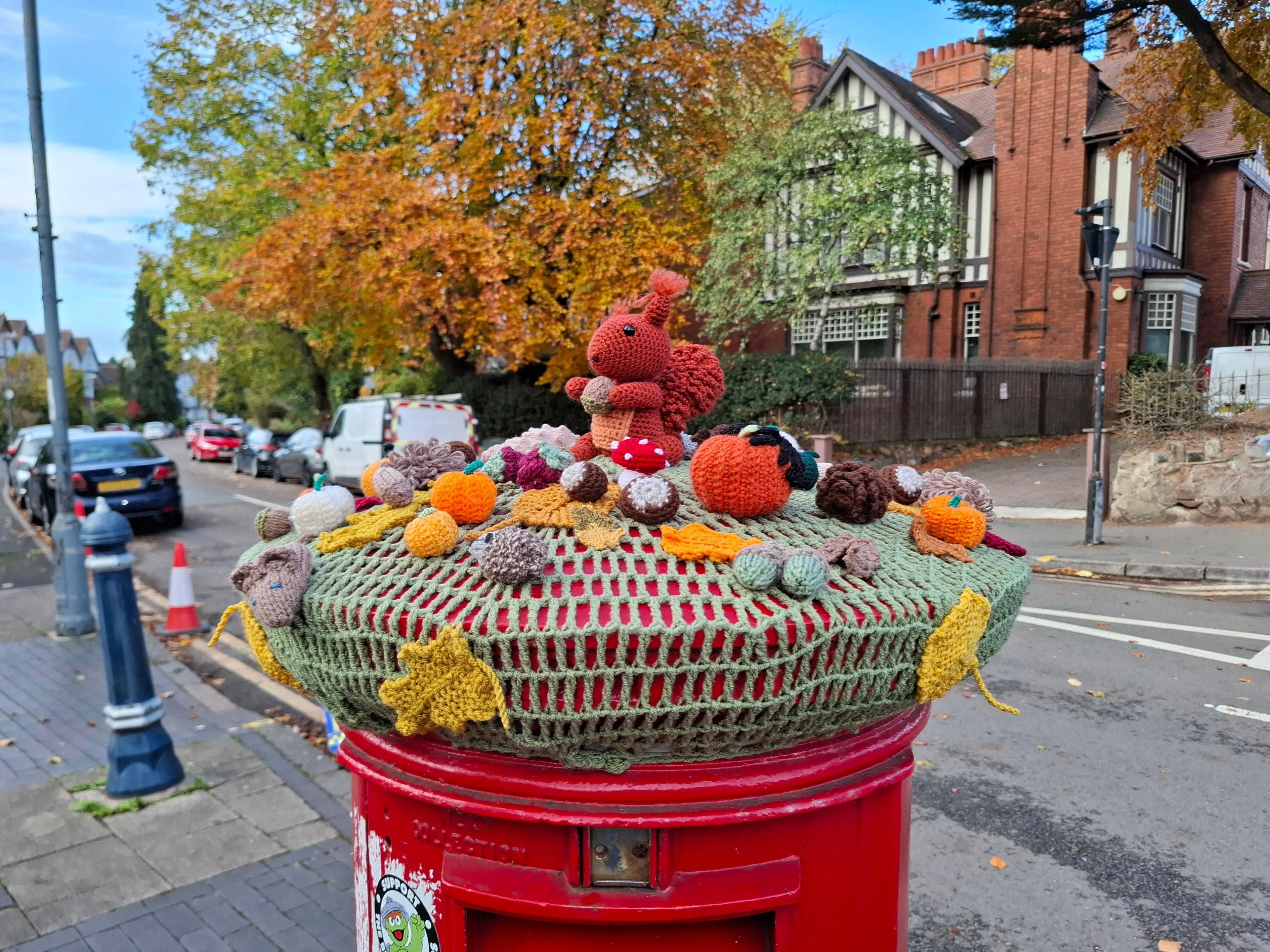 A green knitted base on top of a round red pillar box on a city street. Knitted adornments to the topper include yellow leaves, orange pumpkins, a red mushroom and brown nuts, with a large reddish squirrel at its centre.