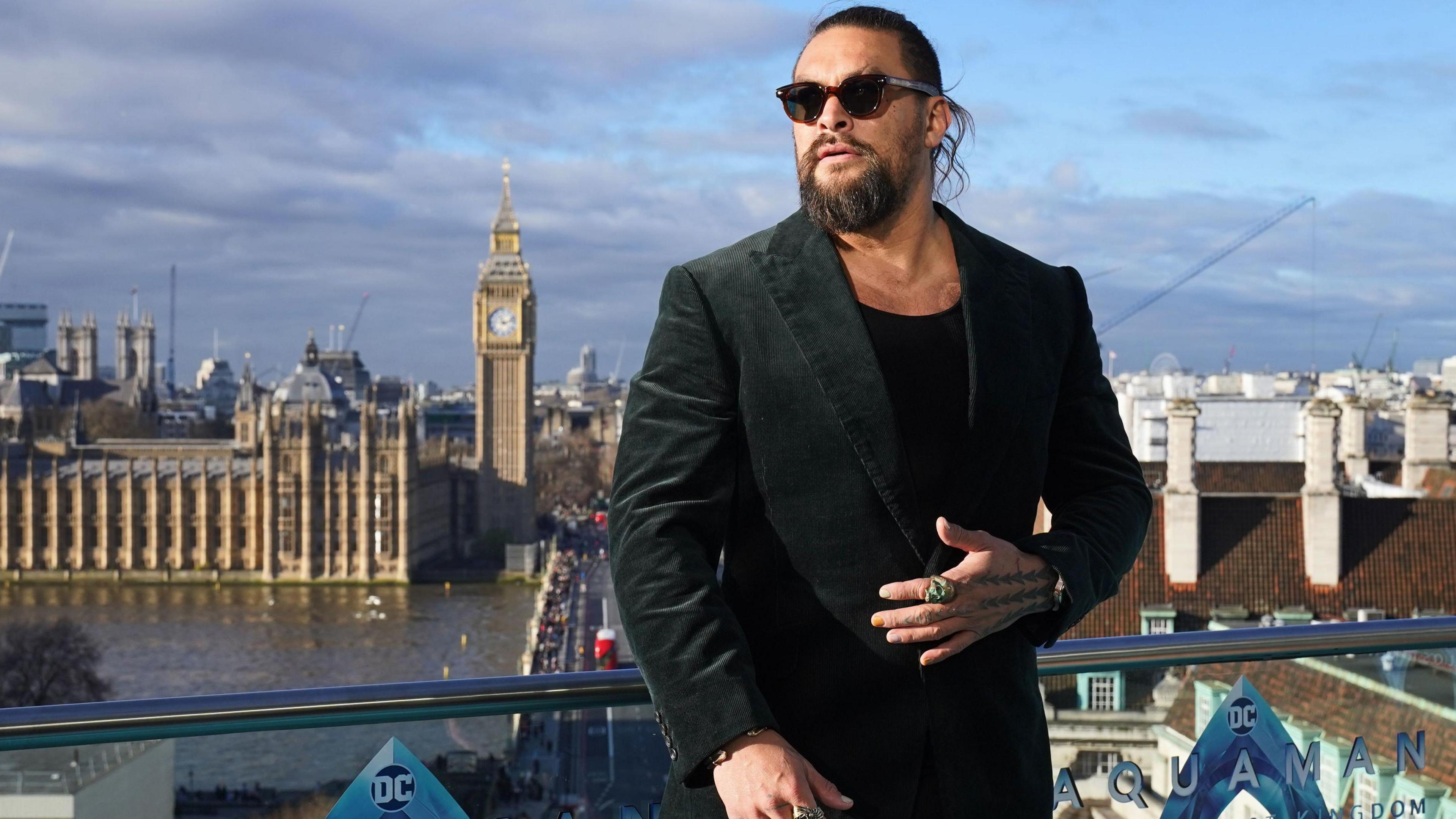 Jason Momoa standing on a balcony on the south side of the River Thames in London. Behind him are County Hall and, across the river, the Houses of Parliament. He is wearing a black suit jacket over a black t-shirt, with his left hand (sporting a giant green ring on his index finger) laid across his midriff. He has brown framed sunglasses and is looking slightly to his right. There are logos for the DC film Aquaman on the glass panels of the balcony.