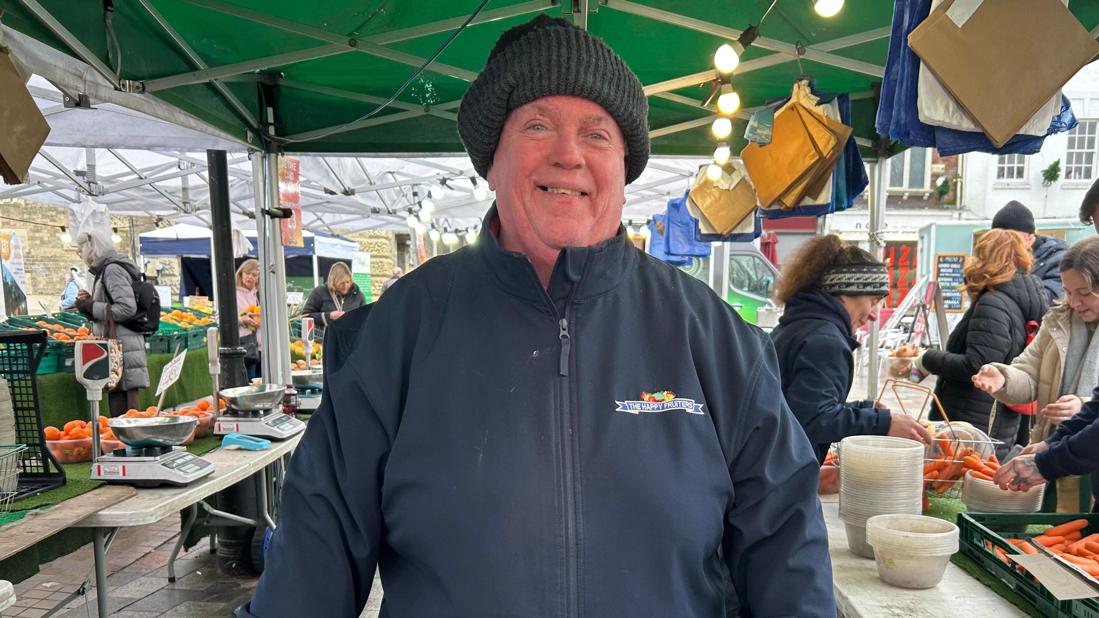 Graham Brown. He is wearing a navy blue jacket with a logo on the chest and a grey knitted hat. He is standing at the market, looking directly at the camera and smiling. 