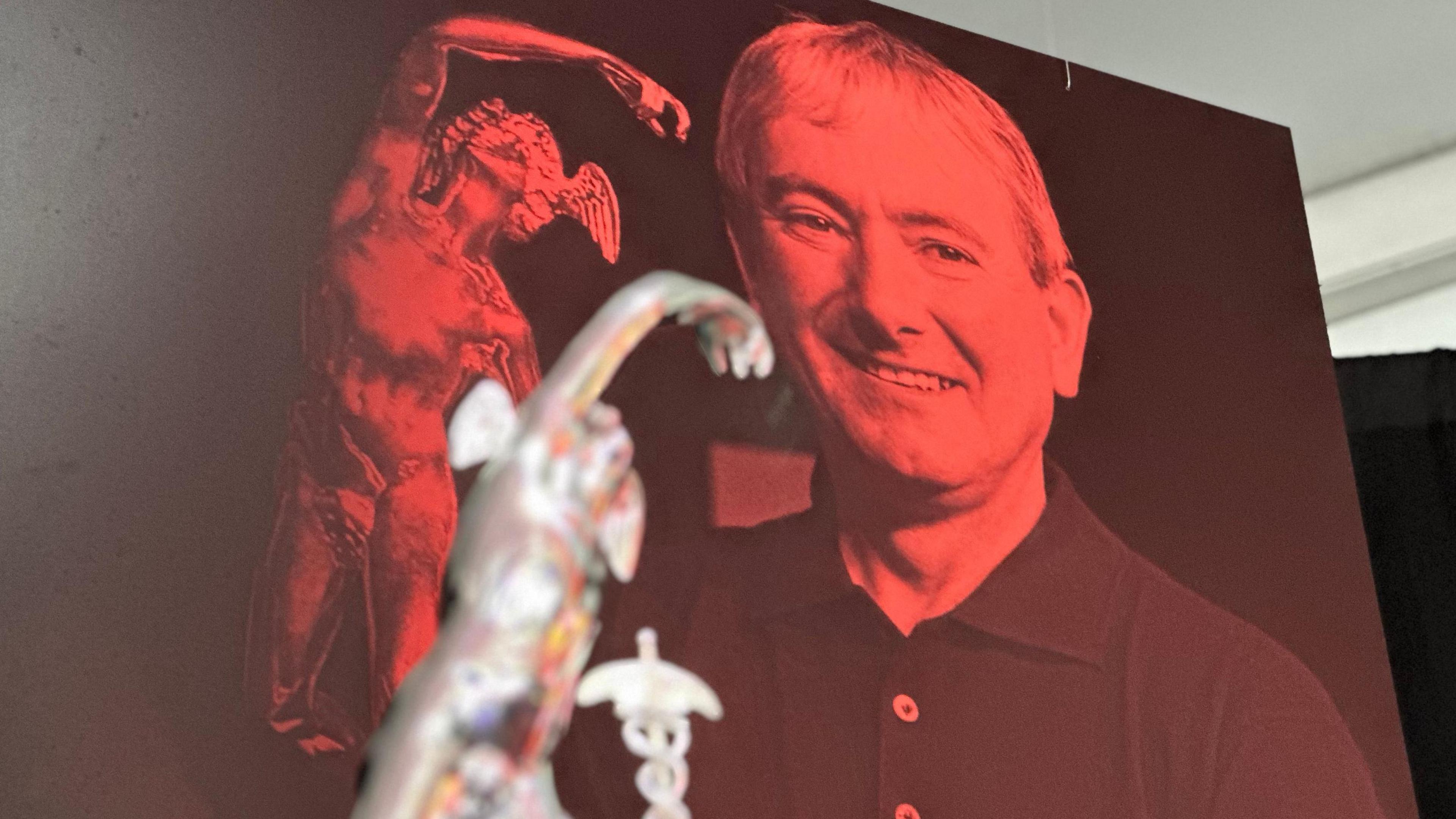 A large red image of Joey Dunlop hangs in the exhibition. He wears a black polo shirt and holds a TT trophy. The top half of a silver TT trophy can be seen in the foreground.