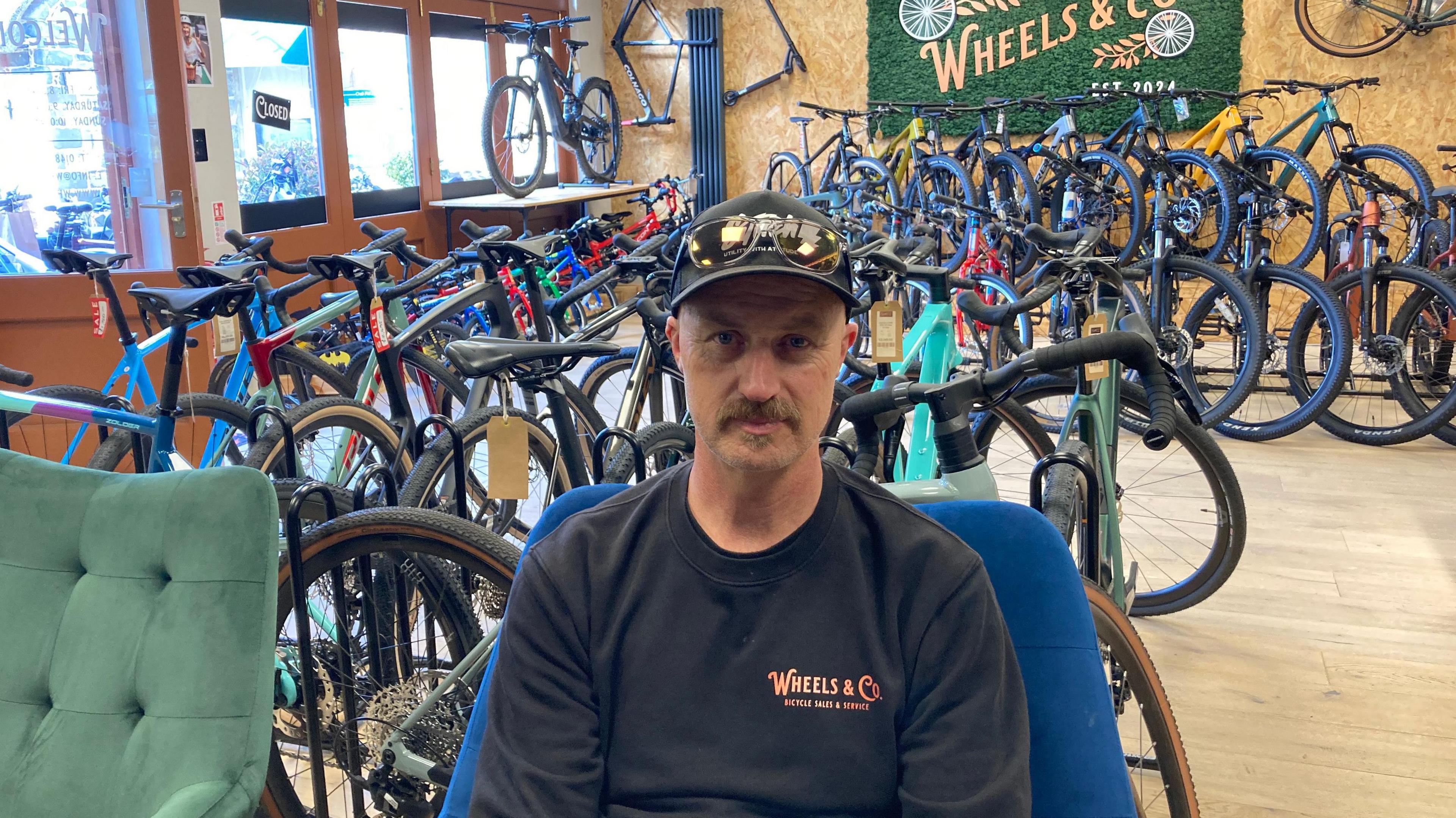A man in a black jumper sitting on a chair in front of bikes in a bike shop