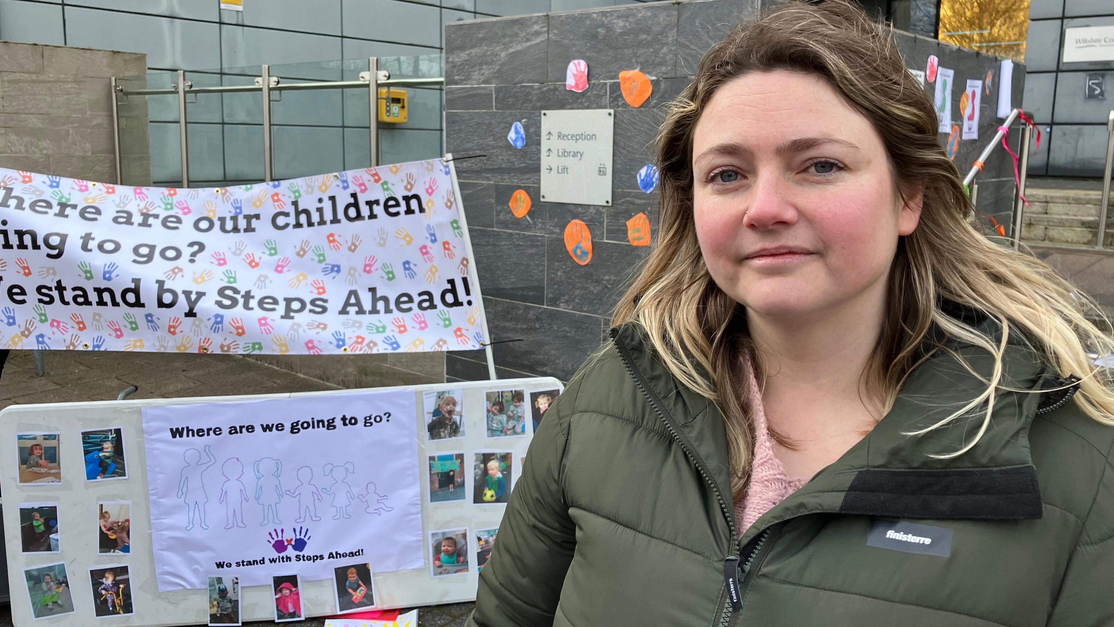 Frankie Simpkins, mother of a child who goes to Steps Ahead Nursery. She is wearing a pink knitted jumper underneath a khaki green padded raincoat. She has mid-length brown hair with bright blonde ends and highlights. Behind her are banners set up advocating for children to stay at the nursery.