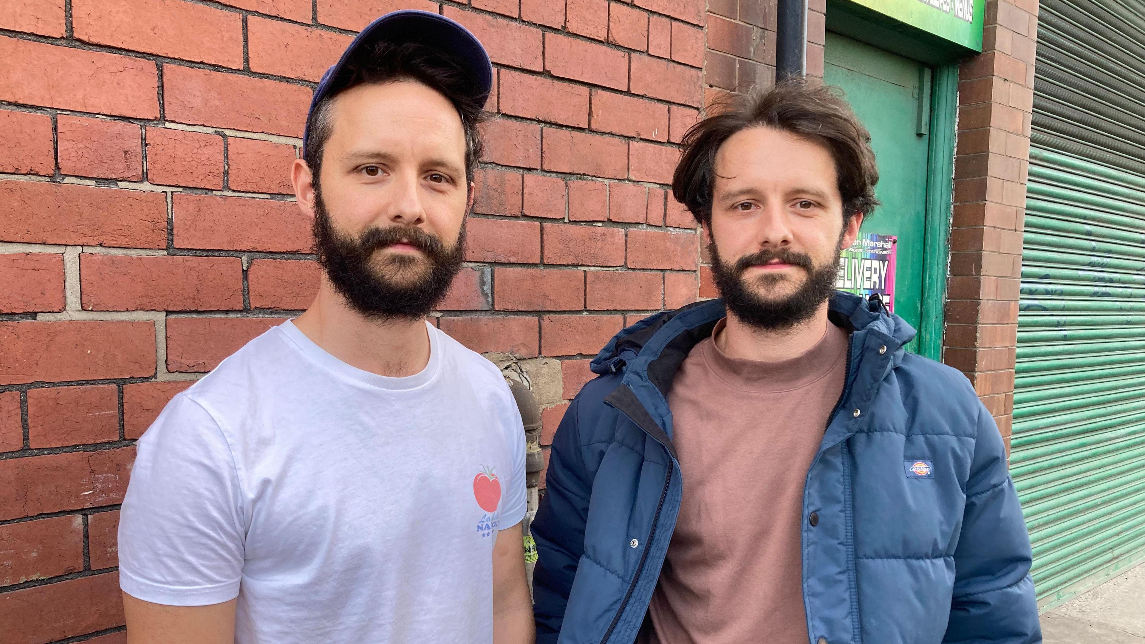 Tom wears a white t-shirt and a cap. He is slightly taller than his brother, who wears a blue coat and a beige t-shirt. Both sport a thick, brown beard.