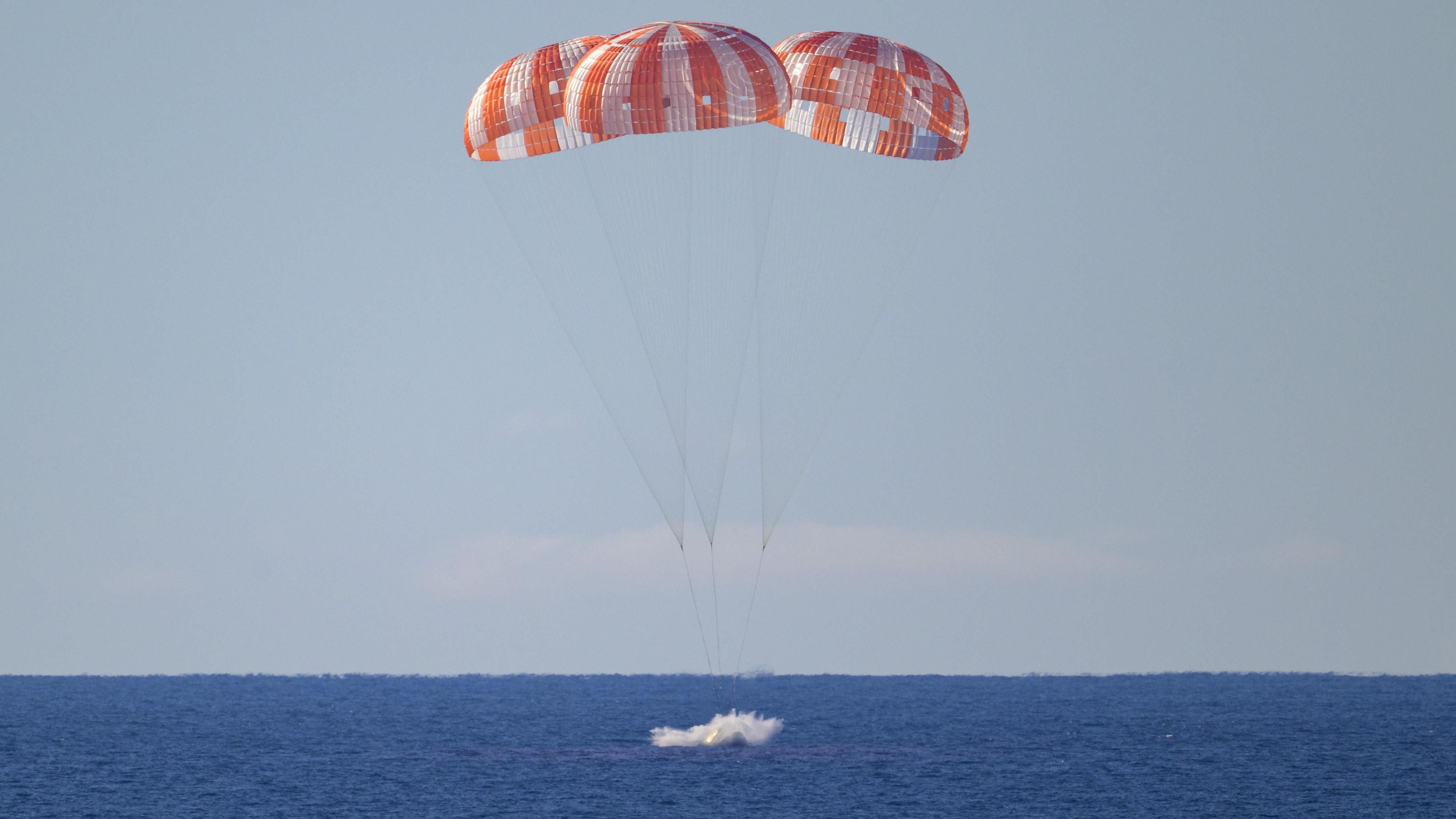Three white and orange parachutes, connected to the Orion capsule float above the sea as the space craft plunges into the water.