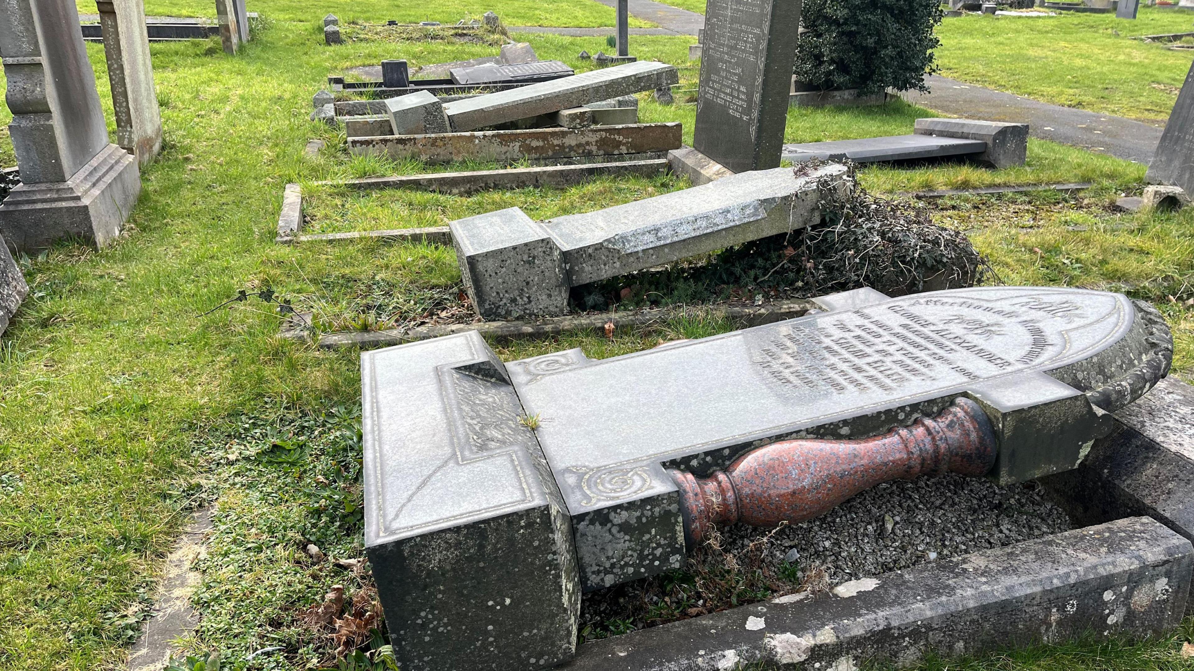 Picture shows several grave headstones uprooted from the ground, knocked over to lay flat. 