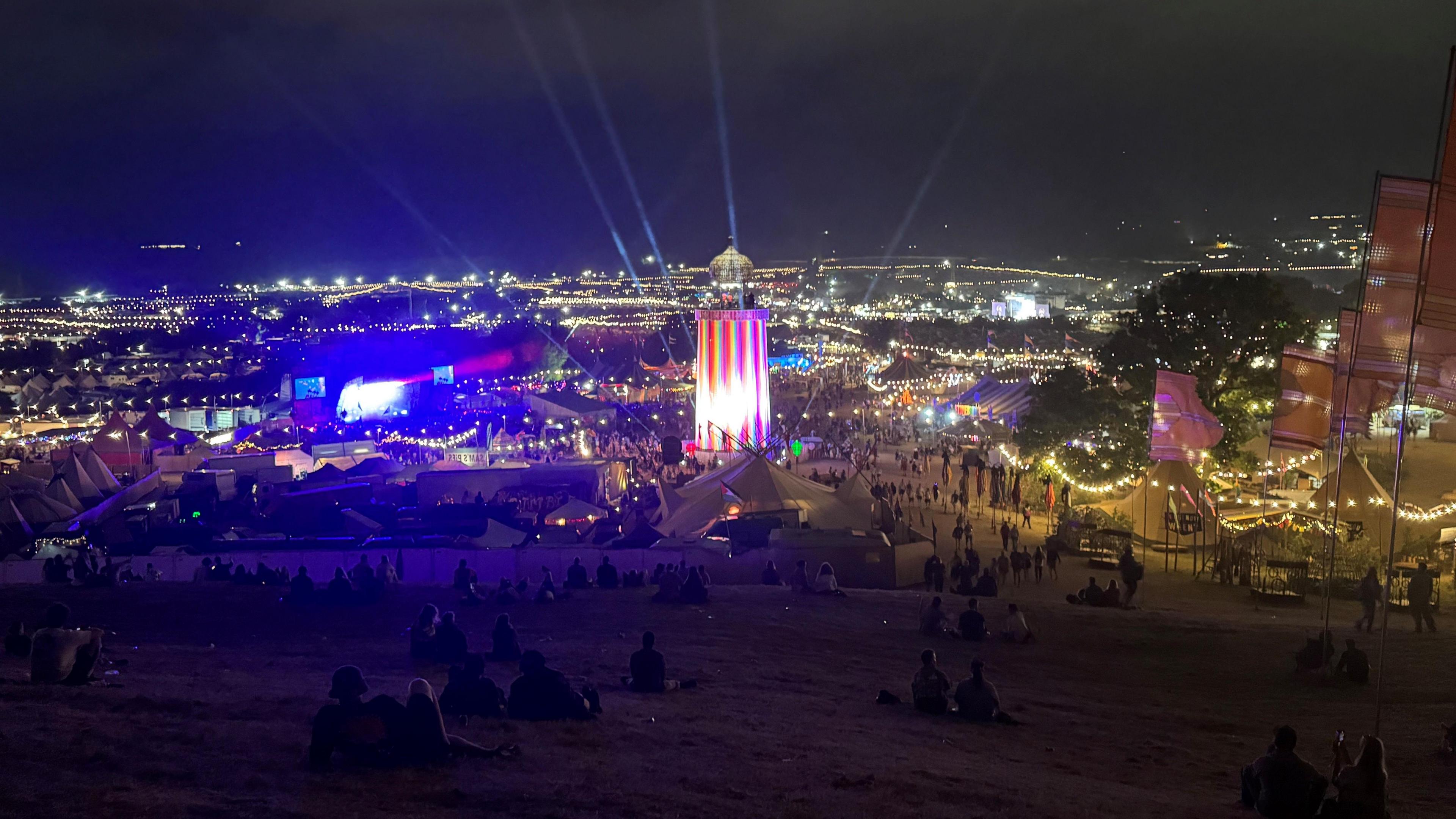 A view across Glastonbury Festival taken at night. There are lights as far as you can see. In the centre is the ribbon tower.