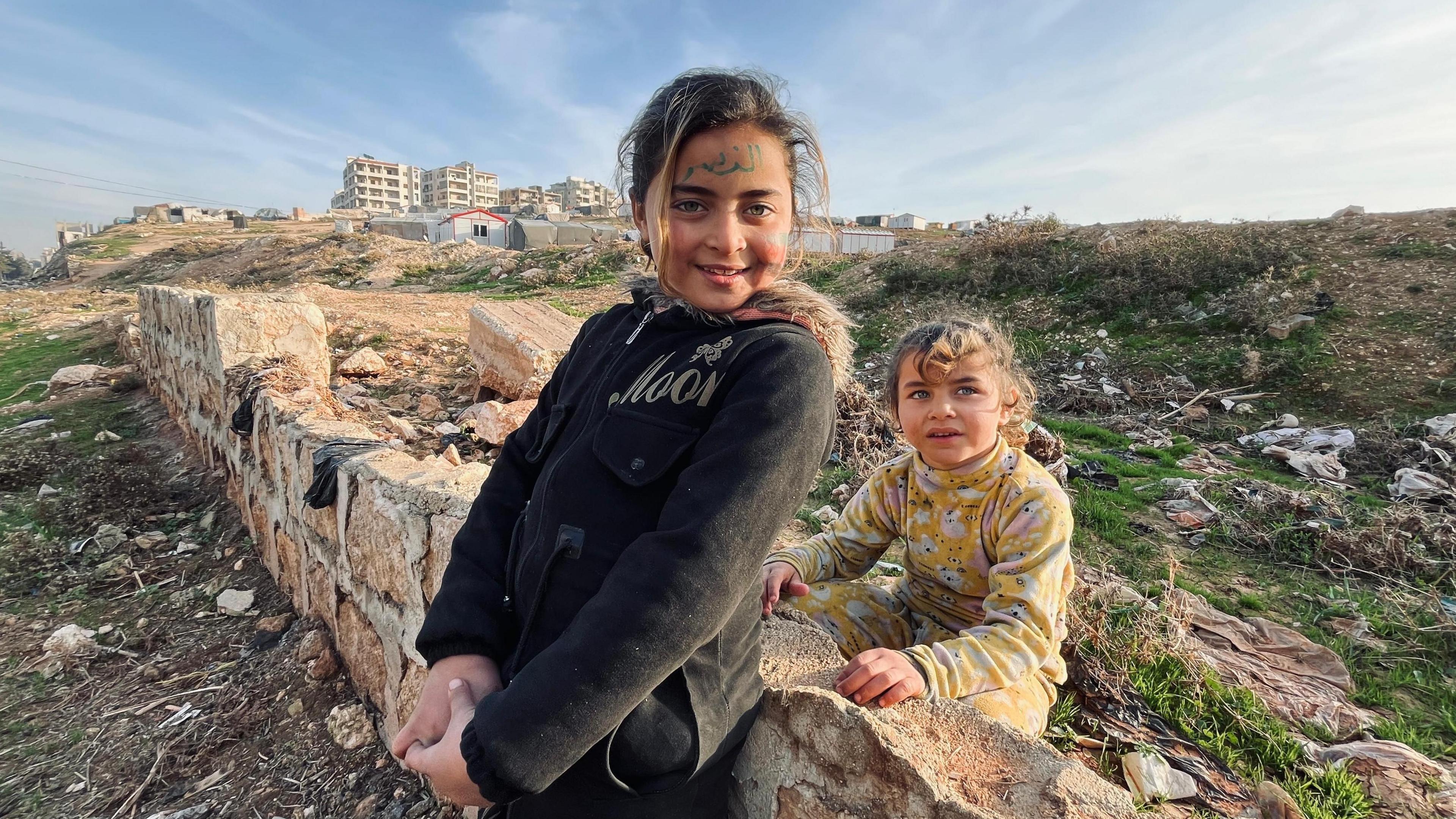 Two young girls stand leaning against a broken wall in a large rubbish pit at an IDP camp in Idlib