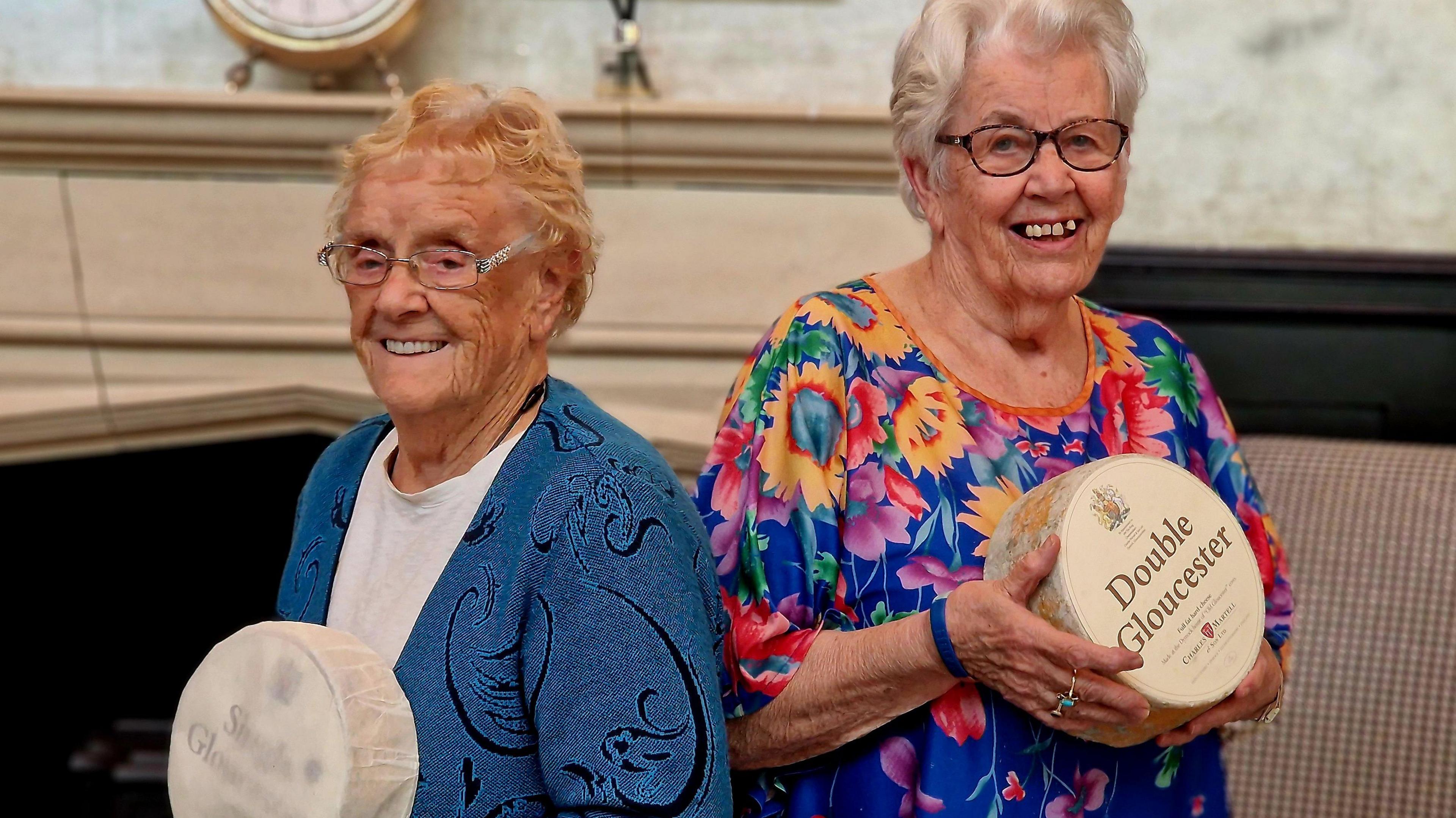 Two women stand side by side holding large wheels of cheese. One holds a wheel labelled "Single Gloucester" and wears a blue cardigan; the other holds a wheel labelled "Double Gloucester" and wears a floral top.