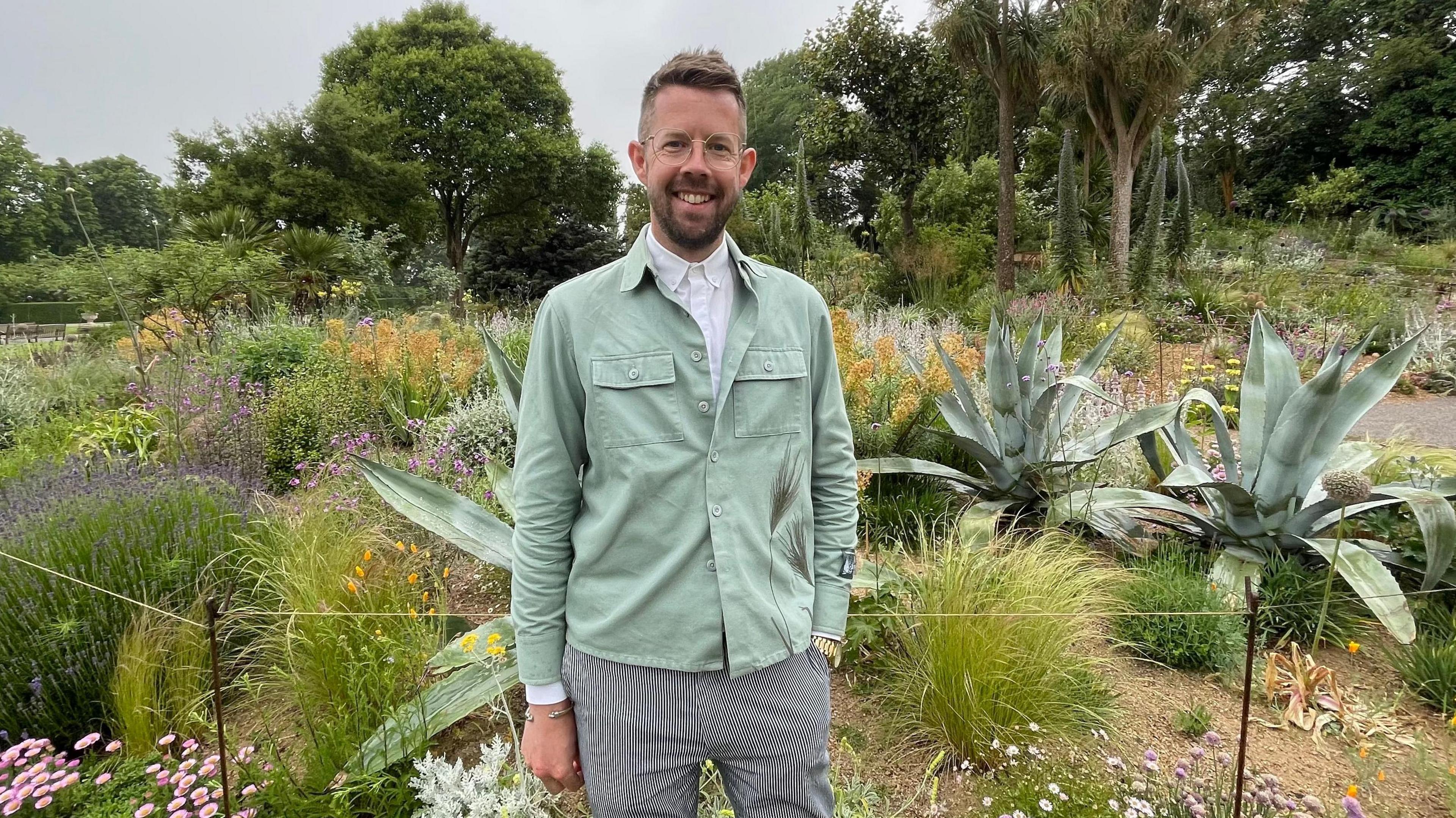 A man wearing a white shirt, mint green jacket and striped trousers stands amongst a garden featuring spiked plants and colourful flowers. He has short cropped brown hair, a beard and is wearing glasses.