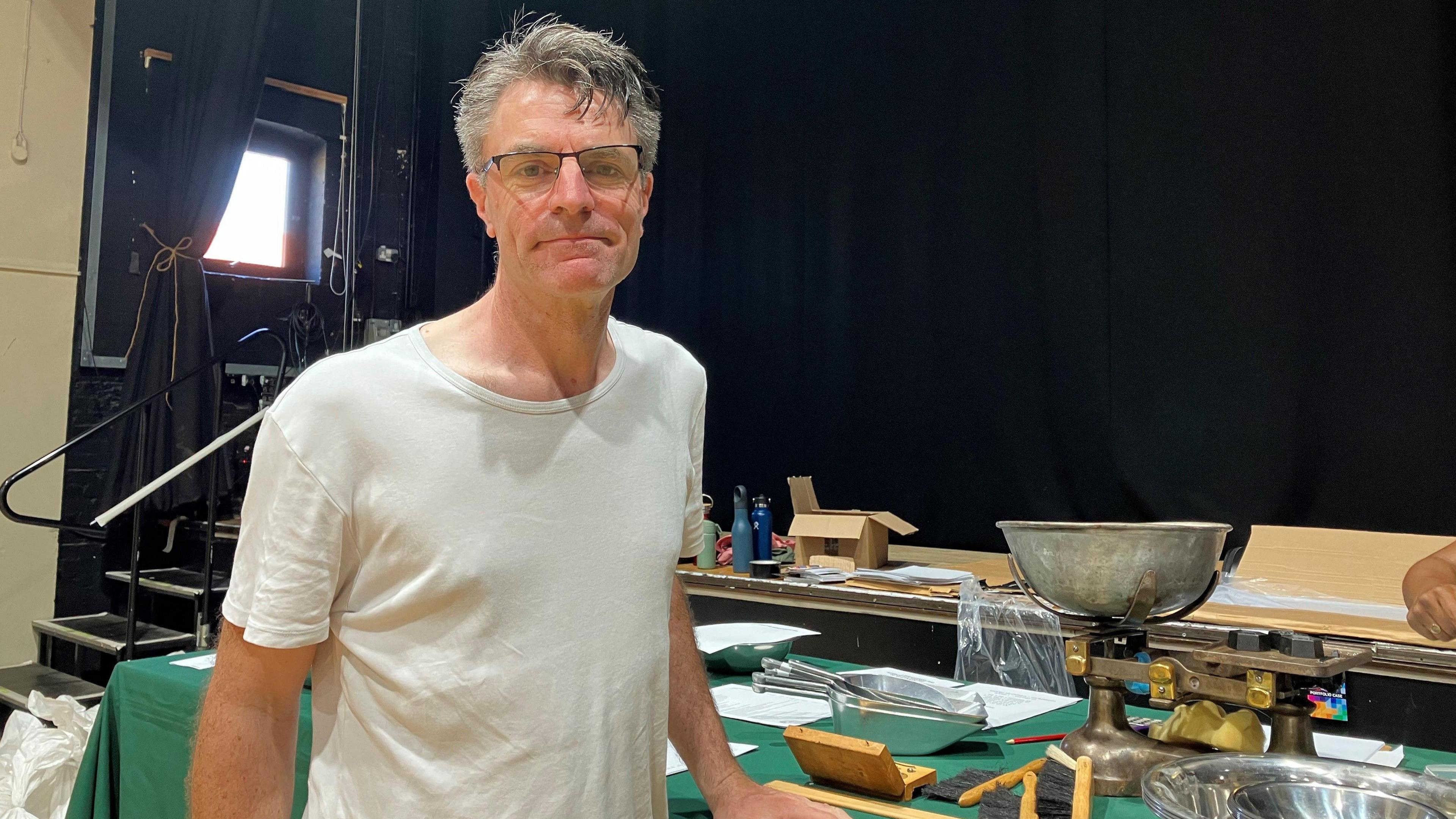 James Yarker looks at the camera and stands in front of a table which has weighing scales on it and various measuring scoops. He is wearing a white t-shirt and has short grey hair.
