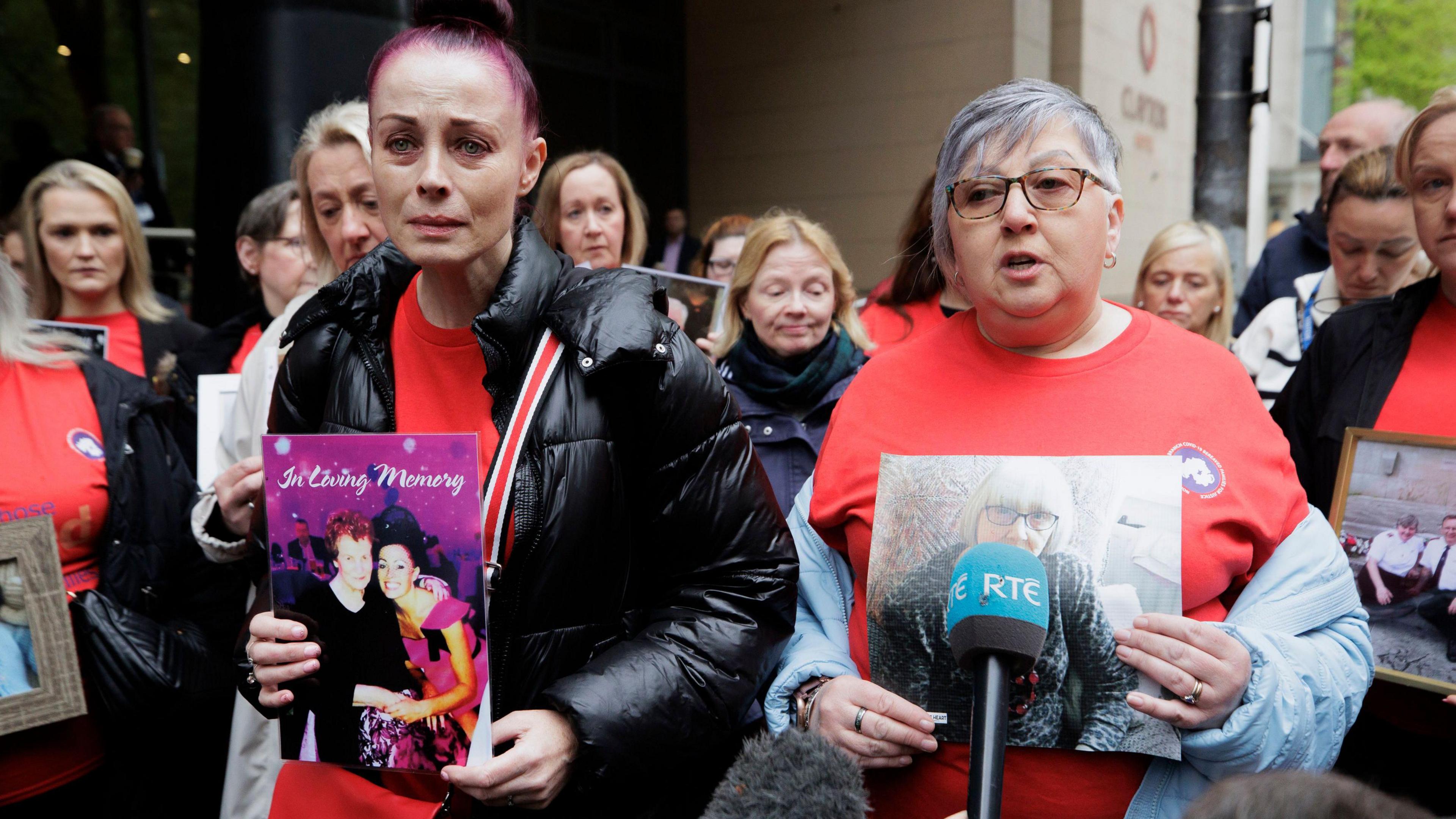 Two women stand at the front of a larger group of people at the infected blood inquiry. They are both holding photos of loved ones. The woman on the left has purple hair and is wearing a black puffy coat over a red T-shirt. The woman on the right has short grey hair and is wearing glasses and a red T-shirt. A teal microphone with RTÉ written on it is in front of her.