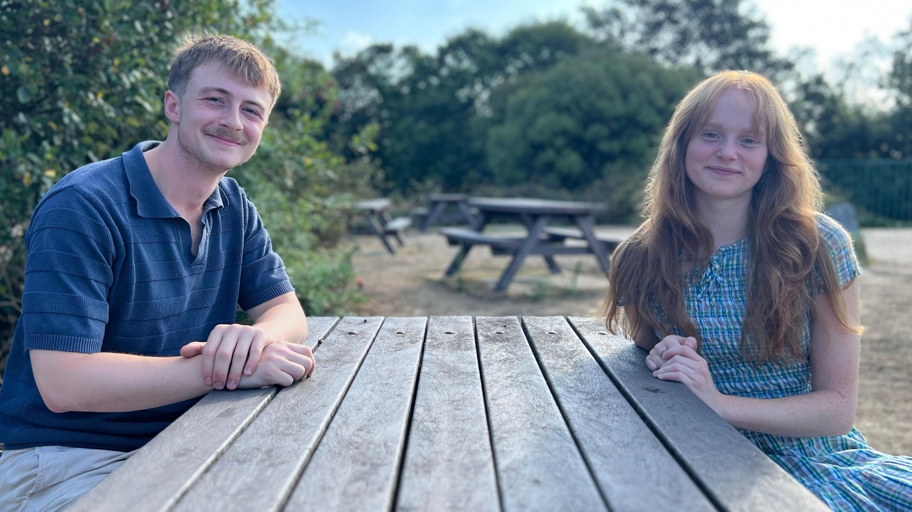 Bill Nicholson is wearing a blue top and sitting at a picnic table opposite Amber Thomson who is wearing a blue dress.