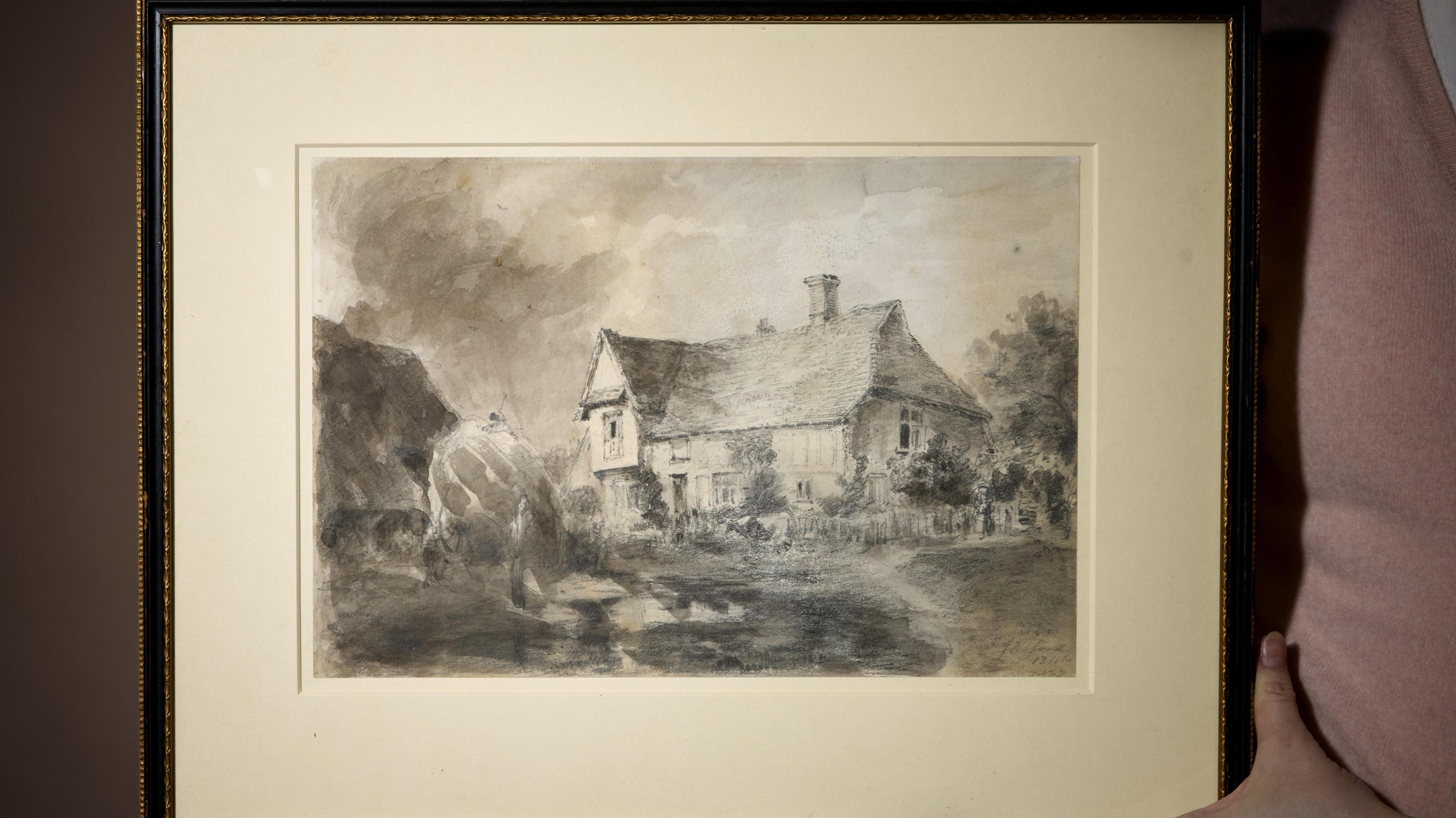 The drawing of a house in the countryside within a black frame. A person wearing a pink top, whose face cannot be seen, holds the frame.