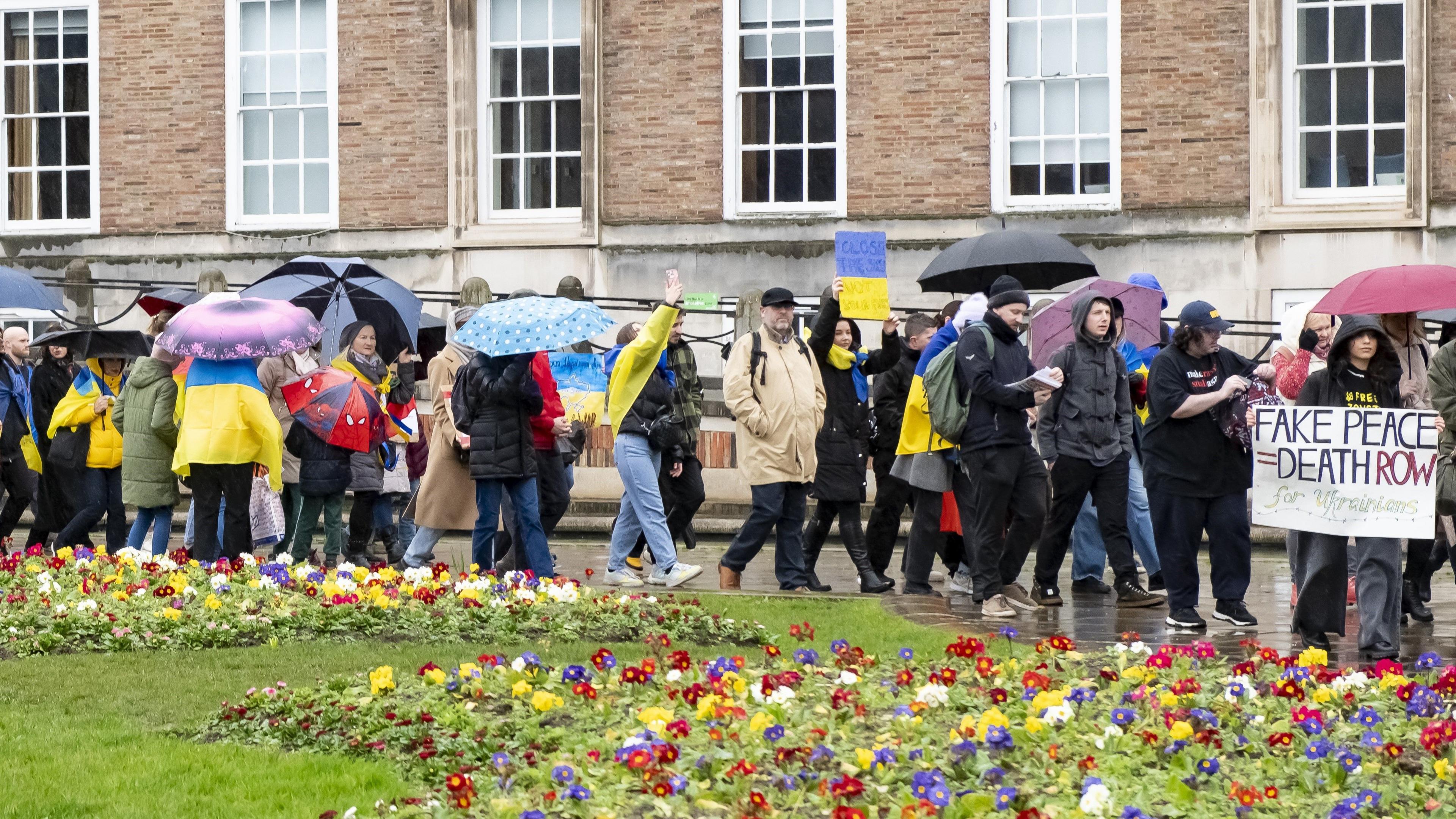 A group of protesters holding signs and Ukrainian flags. They are walking along College Green with City Hall in the background, many with umbrellas. In the foreground there is a bed of colourful flowers and lush green grass.