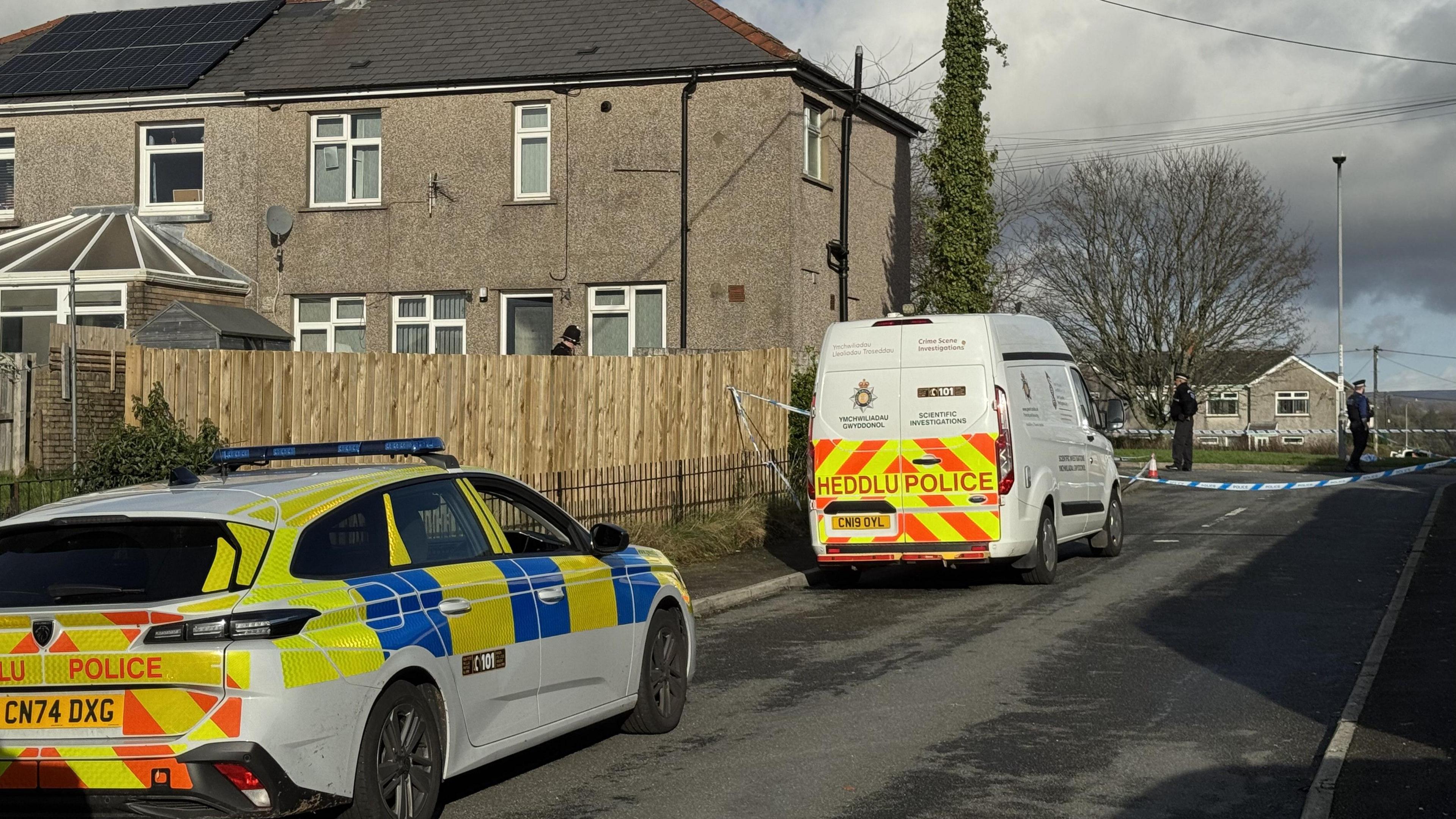 A police car and police van are parked next to the police cordon on a road near to houses, with two police officers behind the cordon.