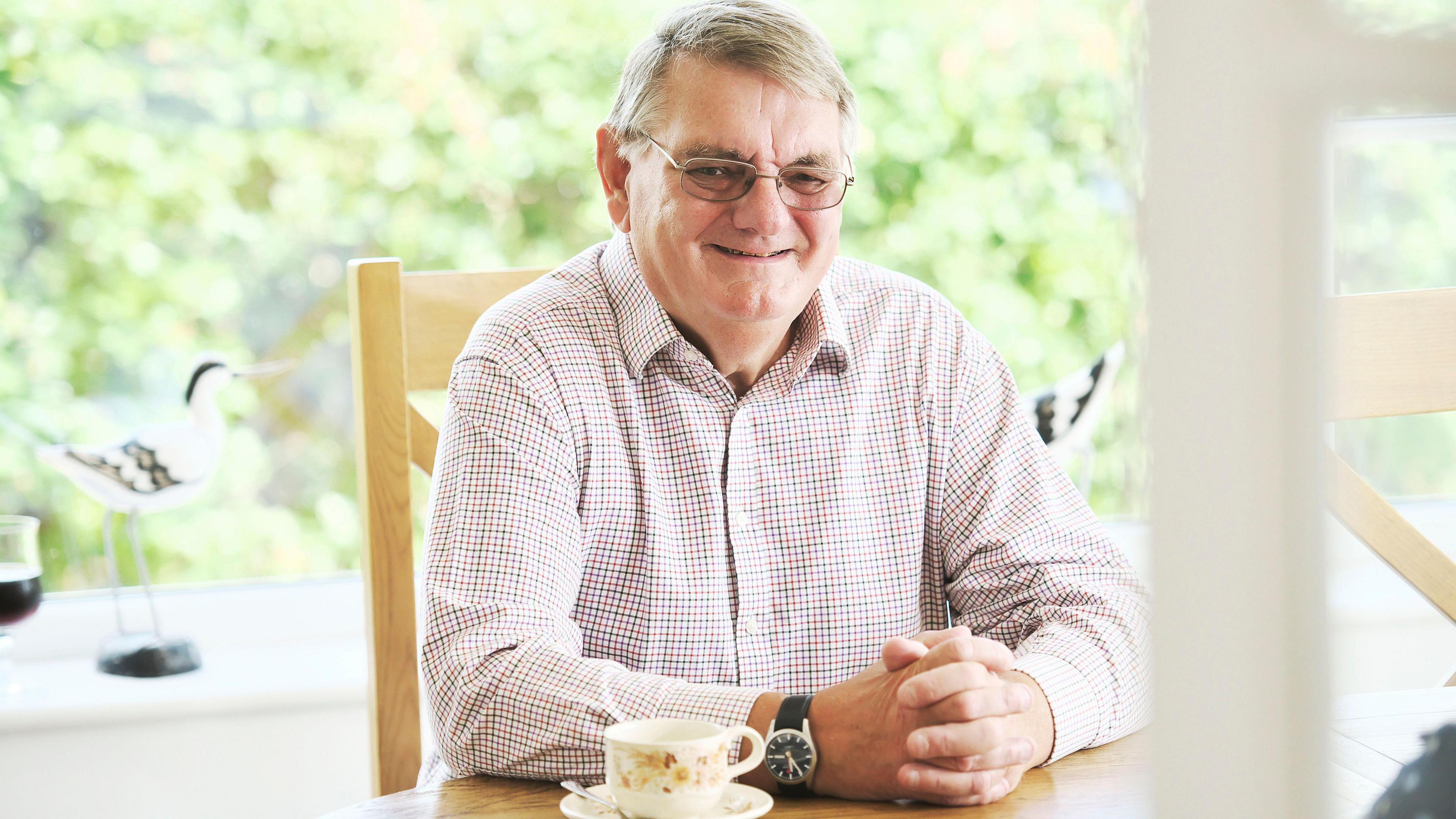 Photo of David oar smiling and sitting at a table. He is a middle-aged man with glasses and a lightly coloured checked shirt.
There is a cup and saucer on the table.